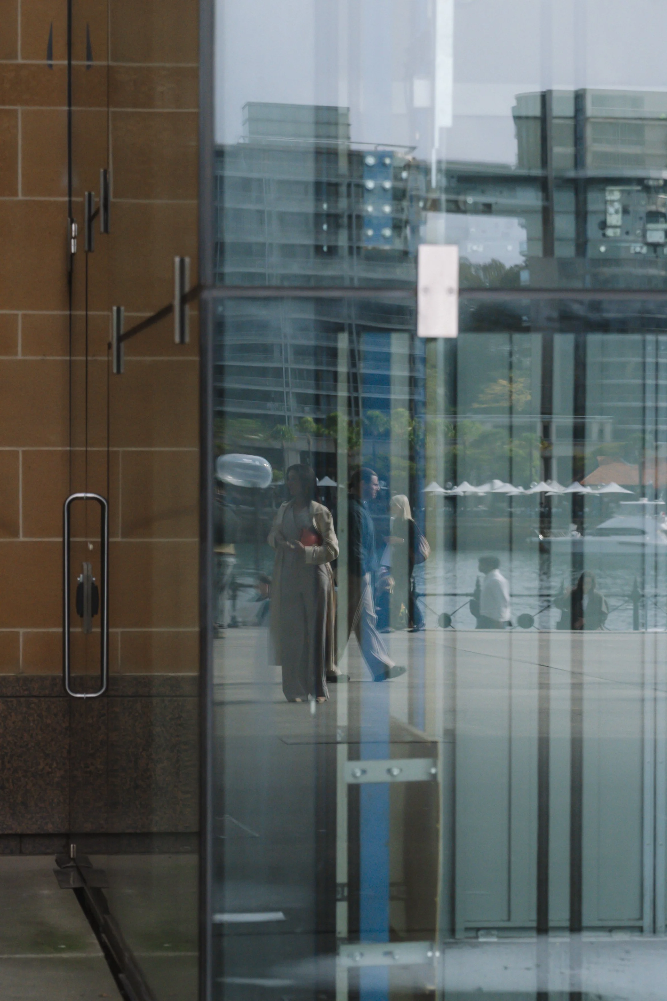 Reflection of people inside a modern building with glass walls and doors, with some individuals walking outside, and a background of other buildings and trees.