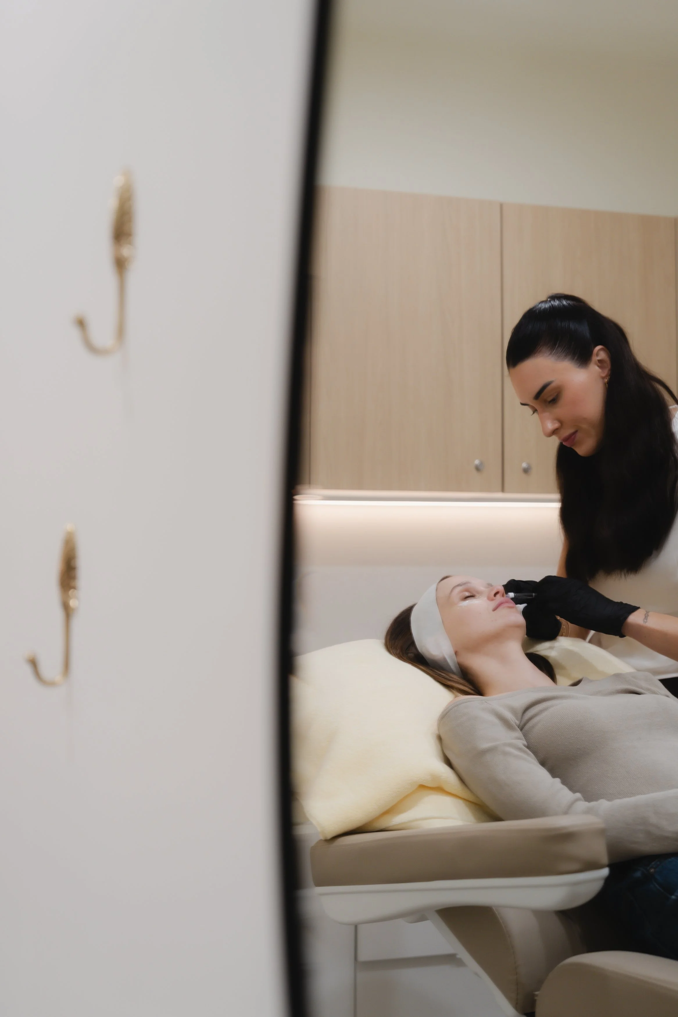 A woman lying on a medical chair with her eyes closed while a healthcare professional administers a facial treatment in a clinical setting.
