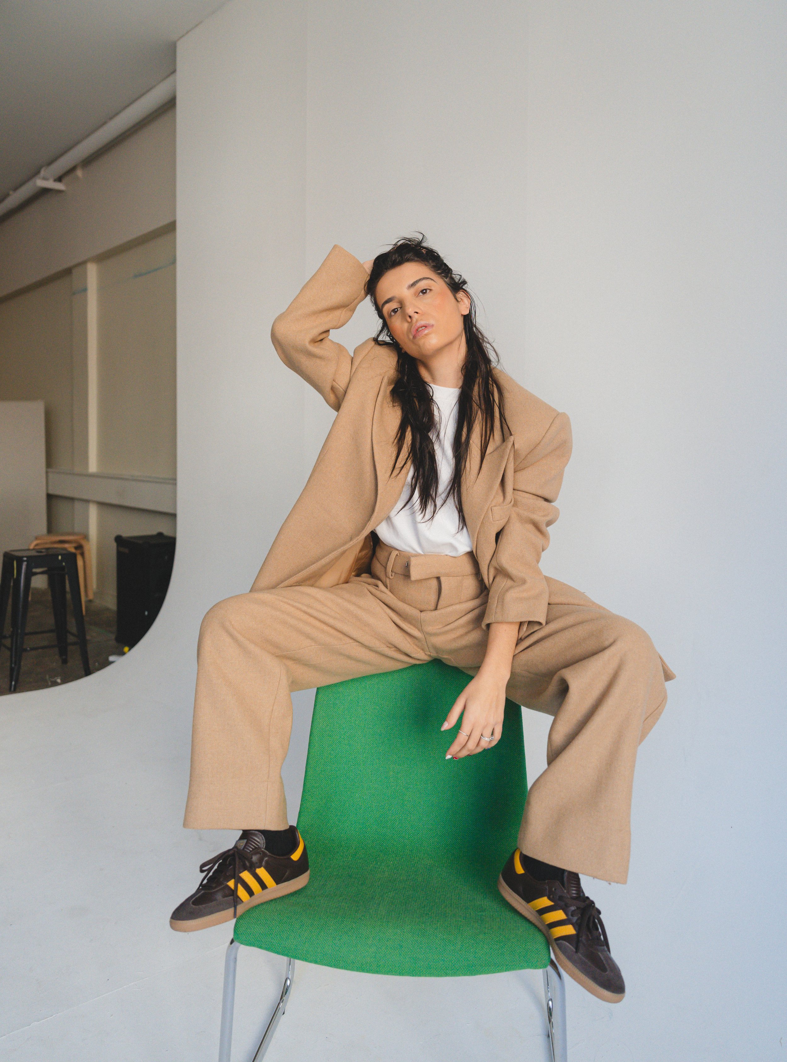 A woman with dark wavy hair wearing a beige suit with a white shirt, sitting on a green chair in a photography studio with a light gray background, holding her head with one hand, and wearing black sneakers with yellow stripes.