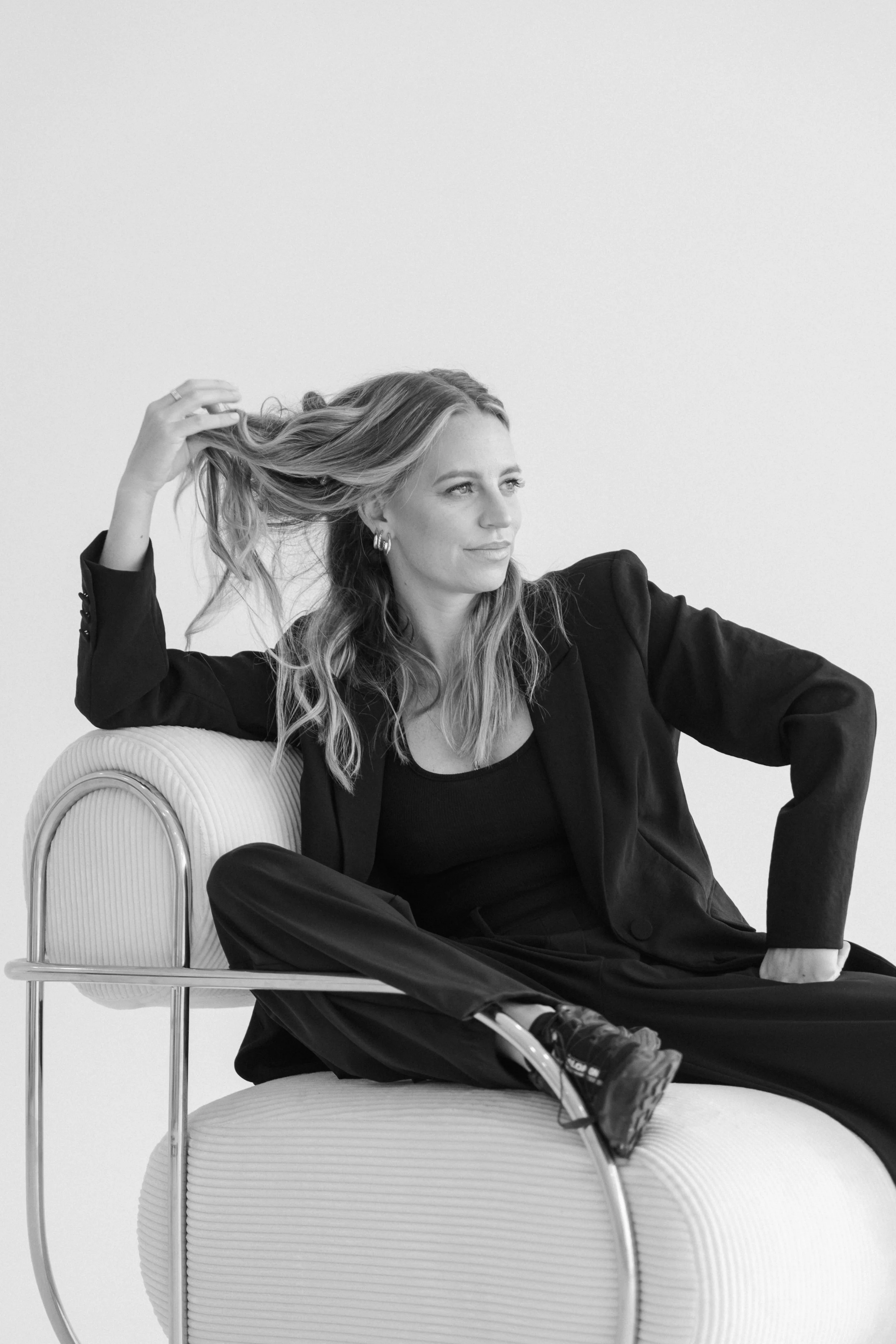A woman with long hair, dressed in a black outfit, sitting on a modern chair, is smiling and looking away while holding her hair. The photo is in black and white.
