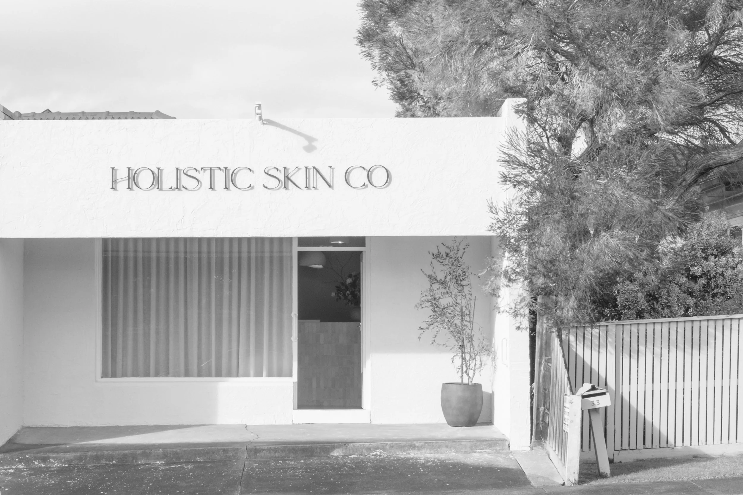 Storefront with the sign 'Holistic Skin Co.' on a white wall, next to windows with curtains. There are trees and plants in front, and a wooden fence on the right side.