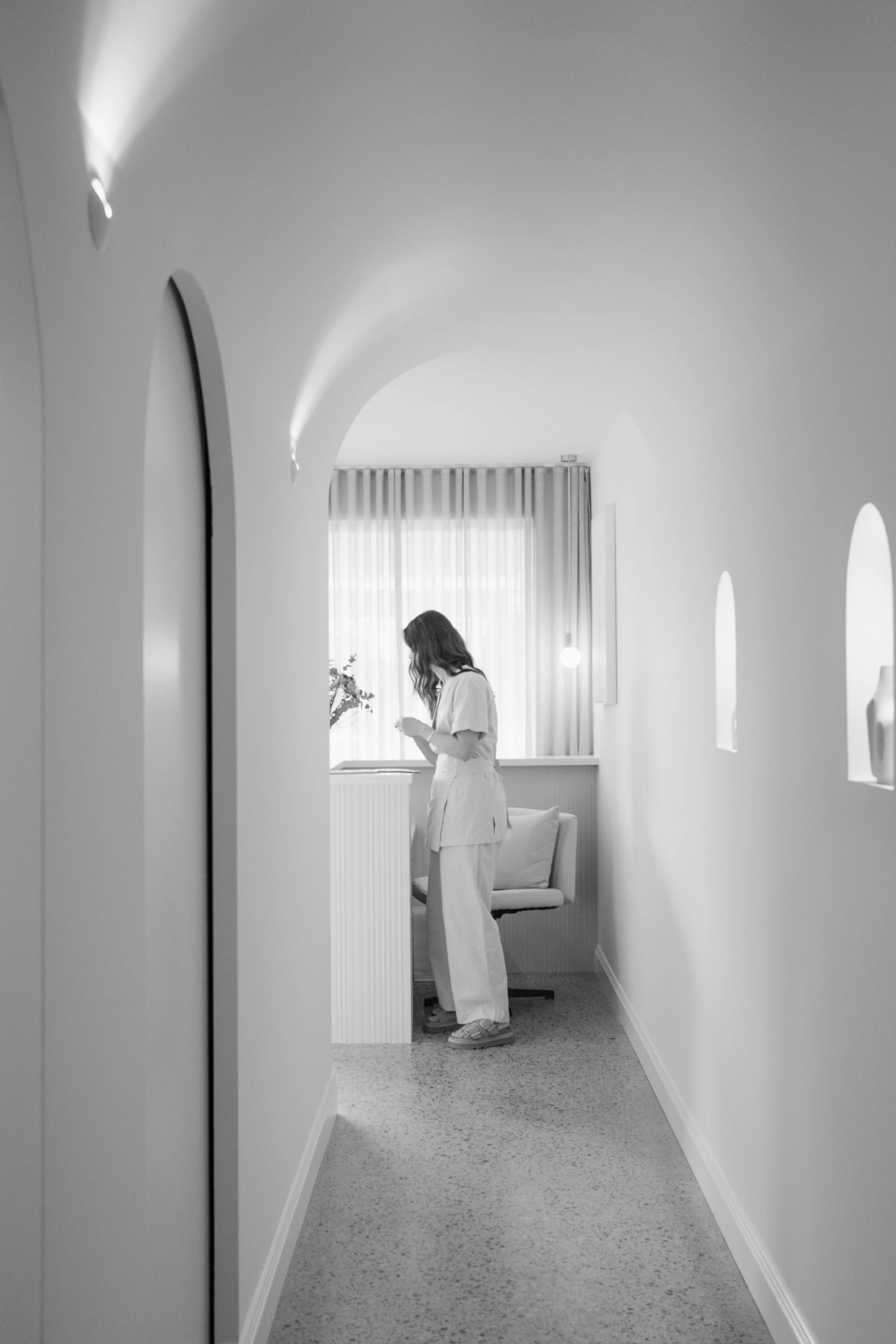 A woman with long hair and casual clothing stands near a window in a well-lit room, with a small sofa against the wall and curtains covering the window.