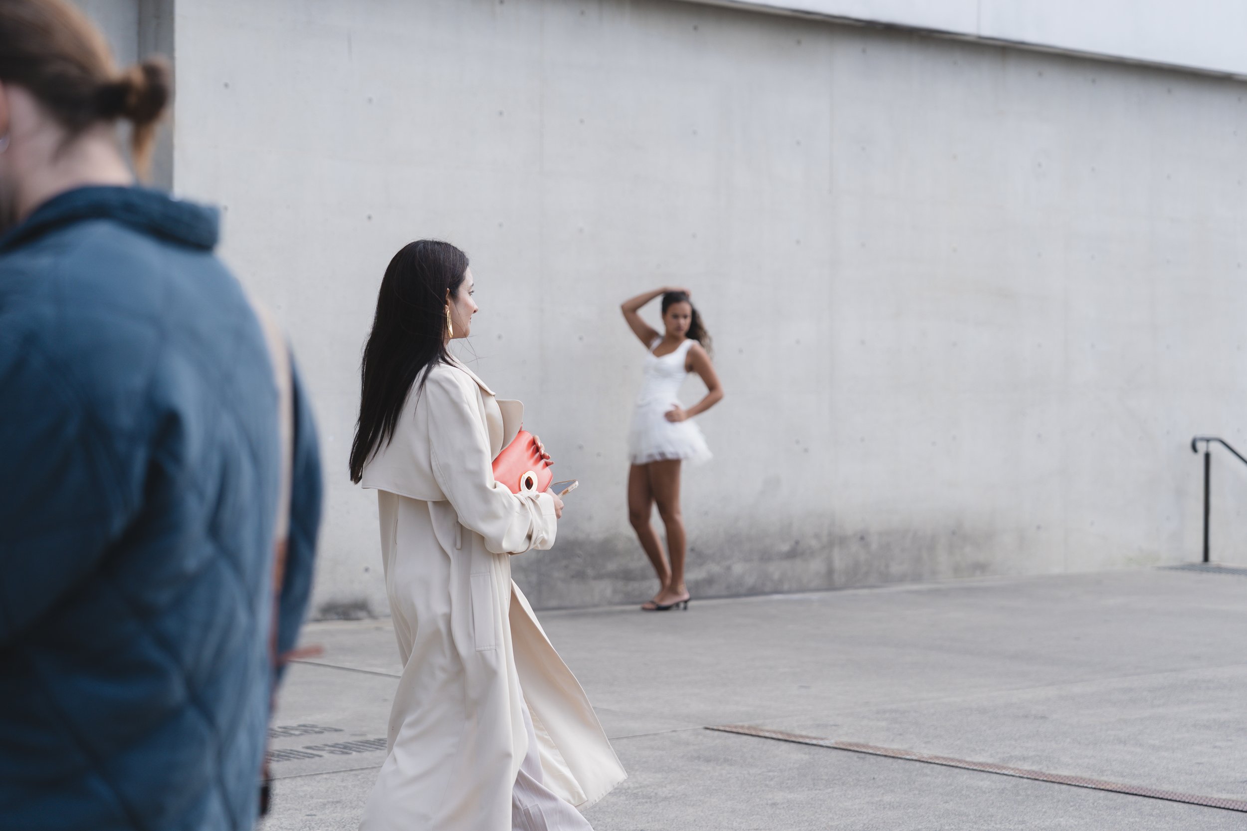 A woman in a white dress walking past a woman in beige coat holding a red purse, with another woman in a white dress posing in front of a large concrete wall.