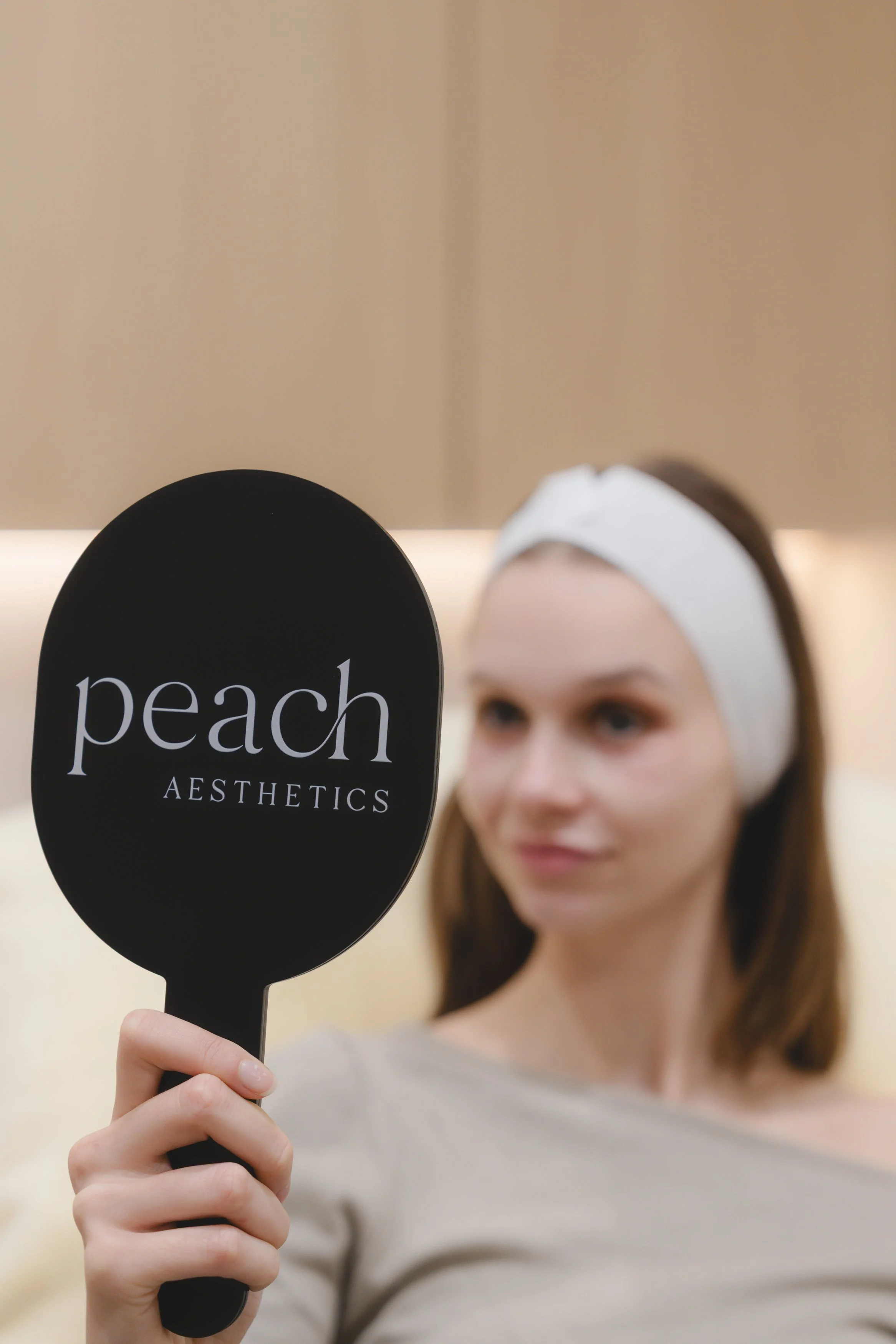 A woman with brown hair, wearing a white headband and white top, holding a black paddle-shaped sign with the words "peach AESTHETICS" printed in white, in an indoor setting with a beige wall background.