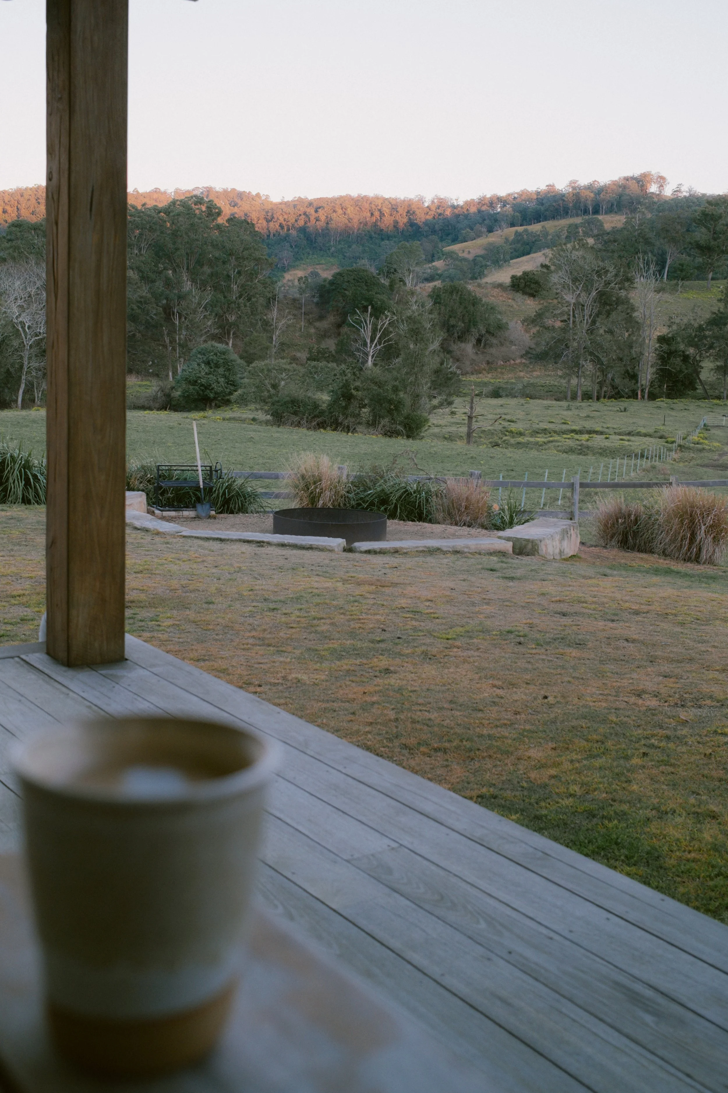 A white cup placed on a wooden deck, with a rural landscape of green fields, trees, and distant hills in the background during daylight.