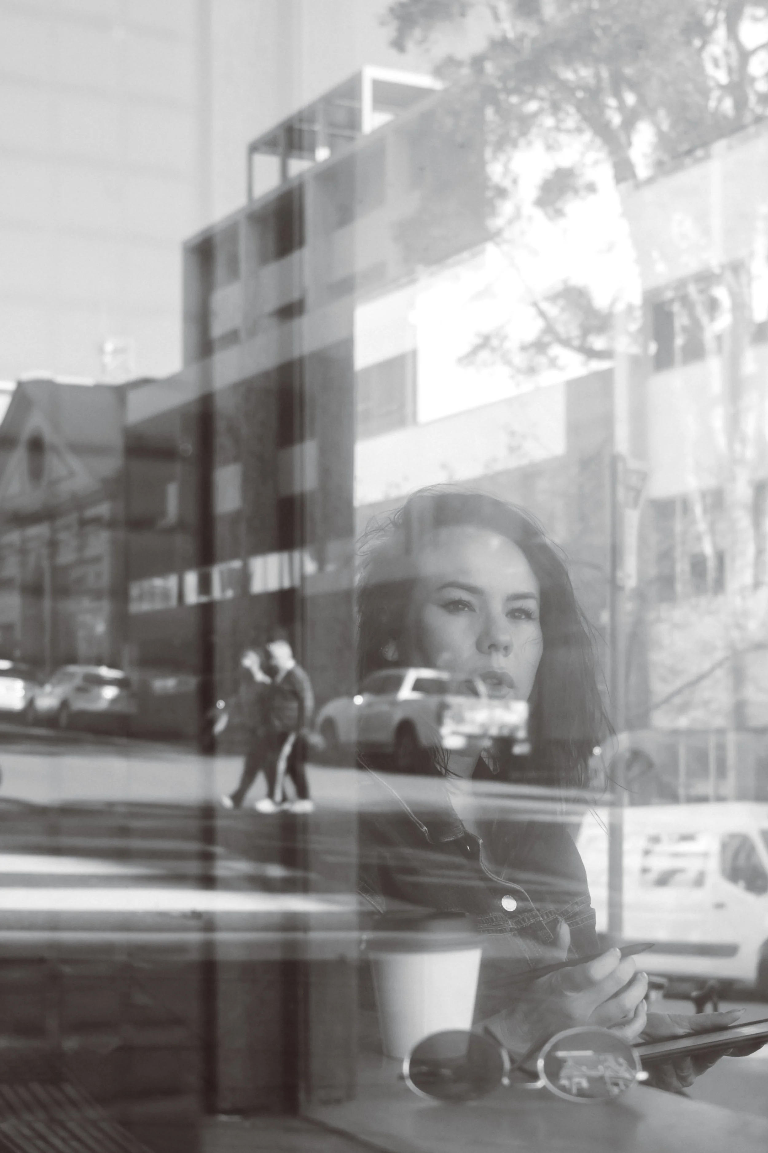 A reflection of a woman taking a selfie in a window with cars and a building outside, with pedestrians walking on the street.