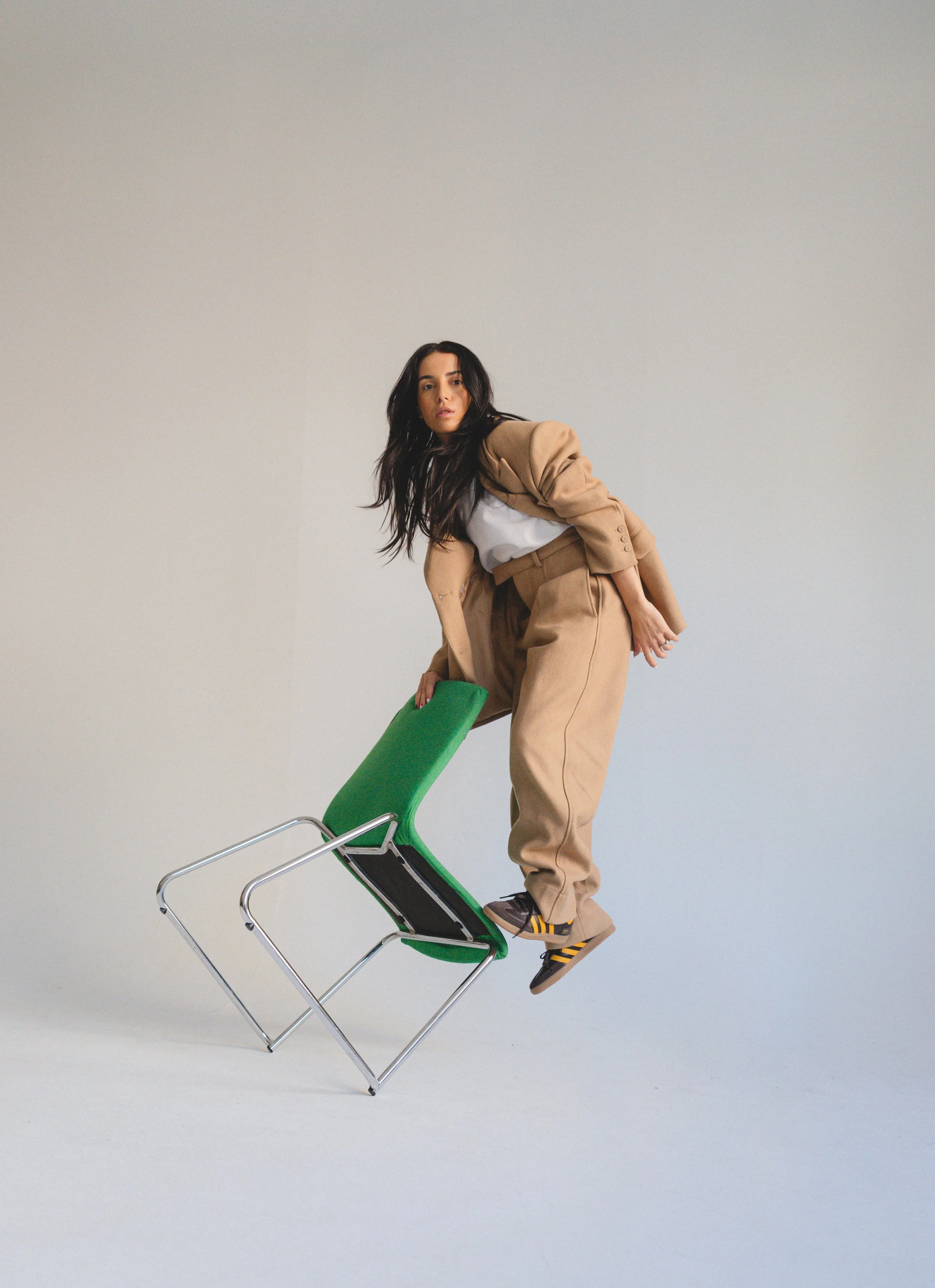 A woman is balancing on a tilted green chair, with one hand resting on the chair and the other hanging down. She is dressed in a beige suit with a white shirt and wears yellow and black striped shoes. The background is plain and light-colored.