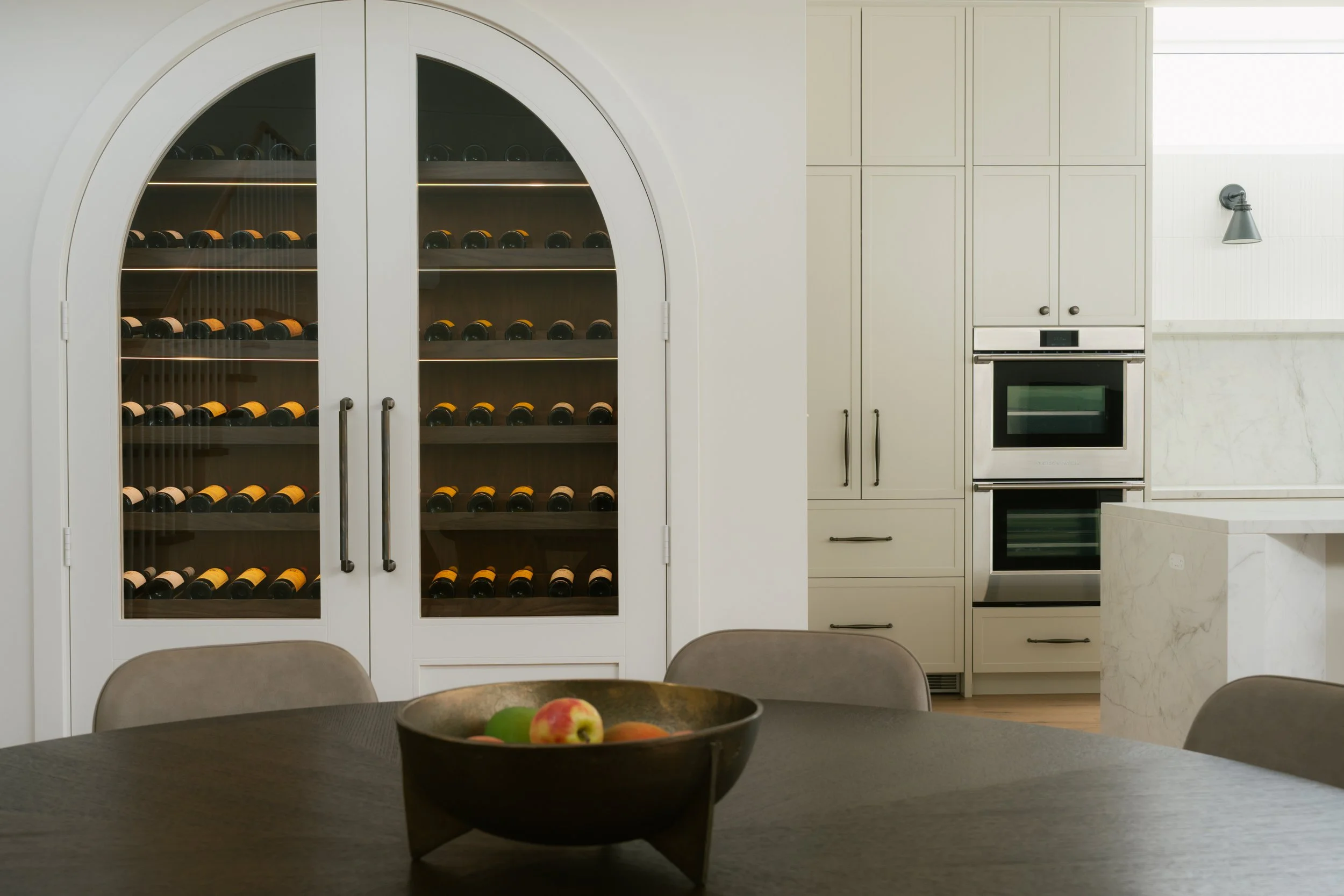 Kitchen with a wine fridge, white cabinets, built-in oven, and a bowl of apples on a black table