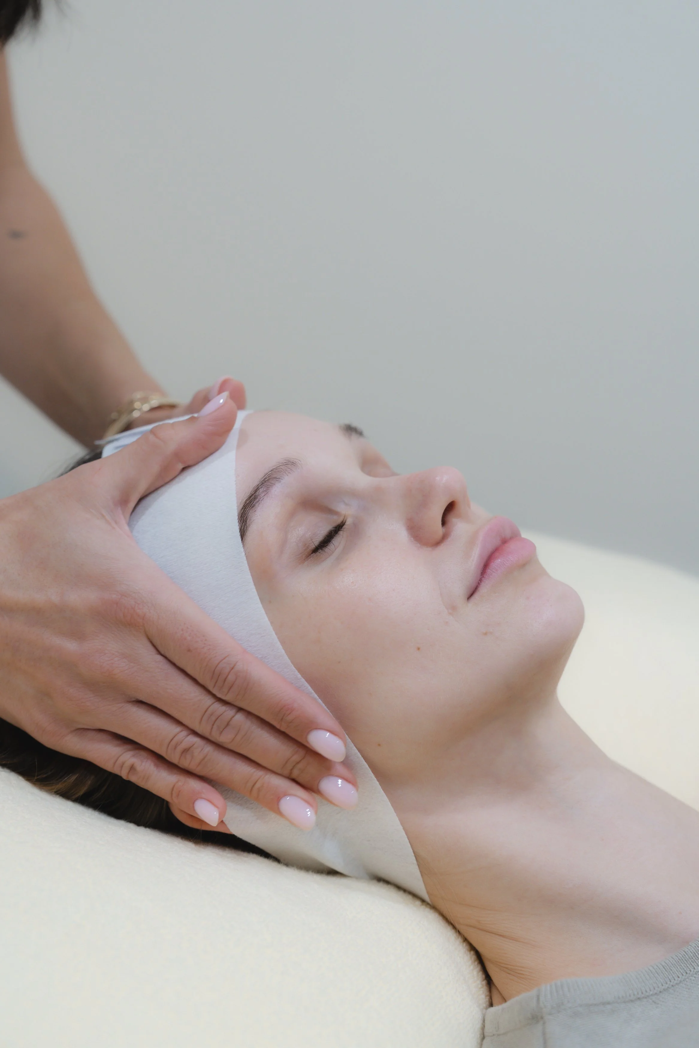 A woman receives a facial massage at a spa, lying down with her eyes closed, while a therapist gently presses her forehead.