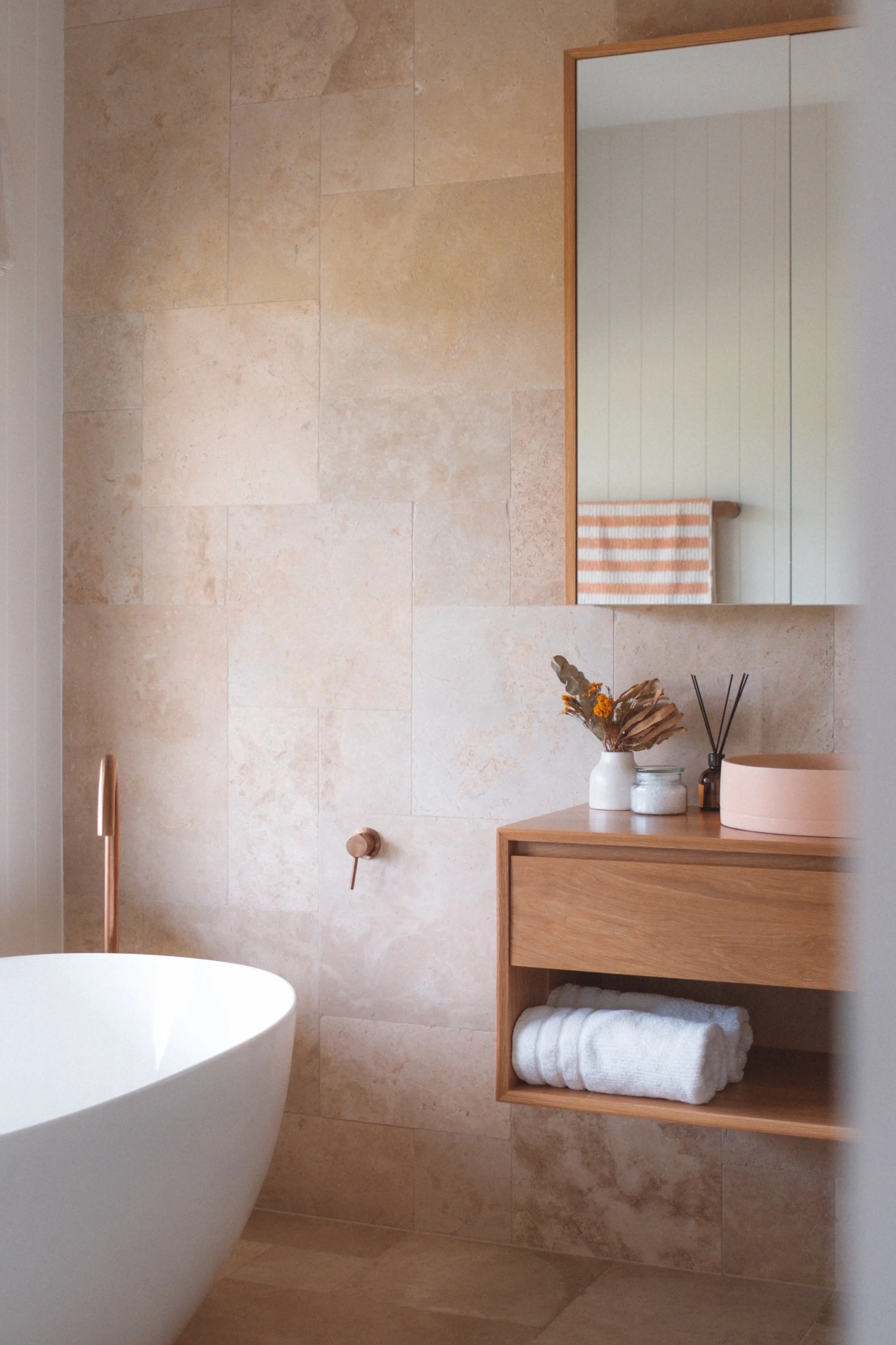 Bathroom with beige tile walls, a large mirror, a wooden shelf with rolled white towels, a vessel sink, and a vase with dried flowers.
