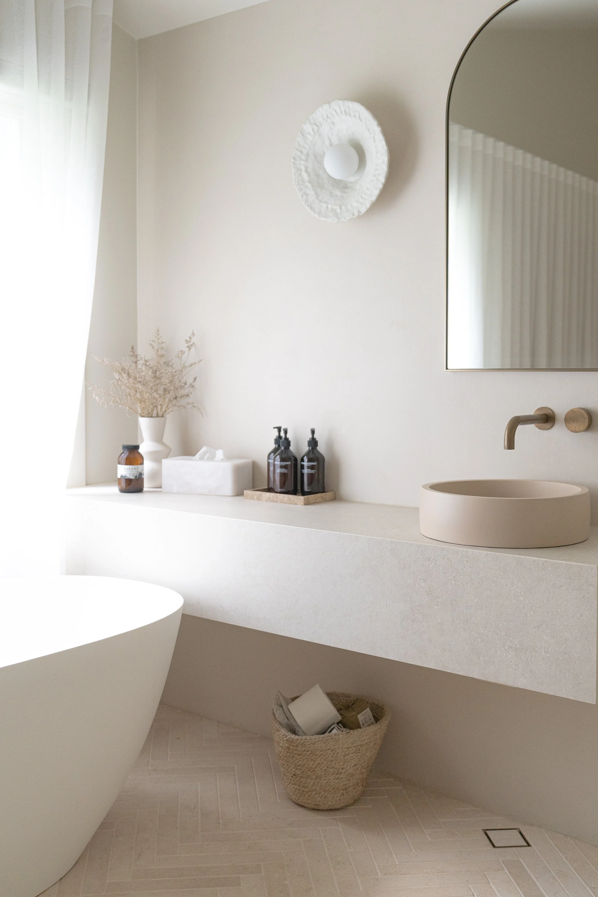 A minimalist bathroom with a white floating vanity, a round vessel sink, a wall-mounted faucet, a large mirror, and decorative elements including vases with dried plants, soap dispensers, a tissue box, and a wicker basket on the floor with rolled tow