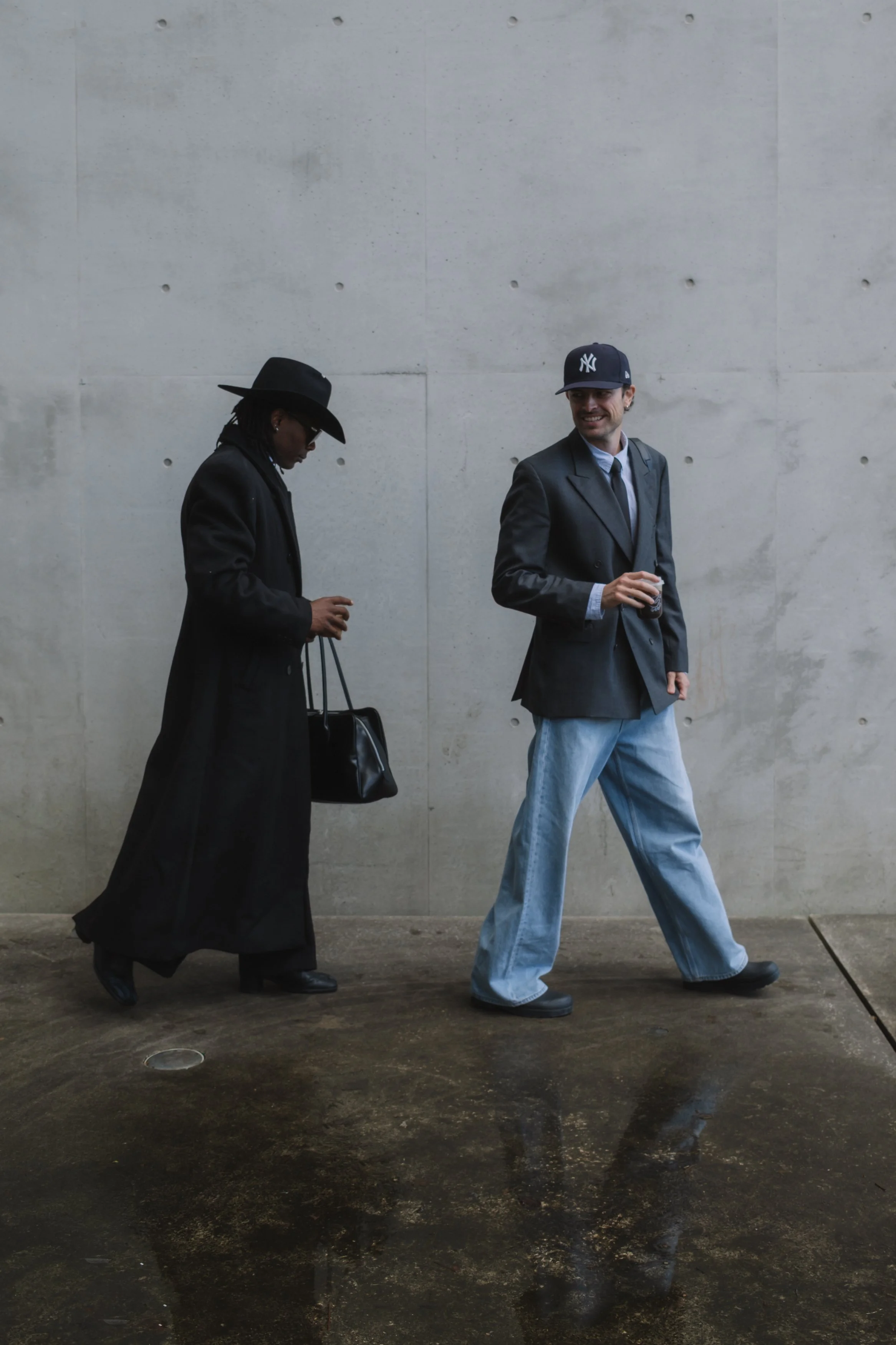 Two men walking past a plain concrete wall. One man is wearing a long black coat, black hat, sunglasses, and carrying a black bag, looking downward. The other man is wearing a dark blazer, a blue baseball cap with the New York Yankees logo, and holding a drink, smiling while looking ahead.