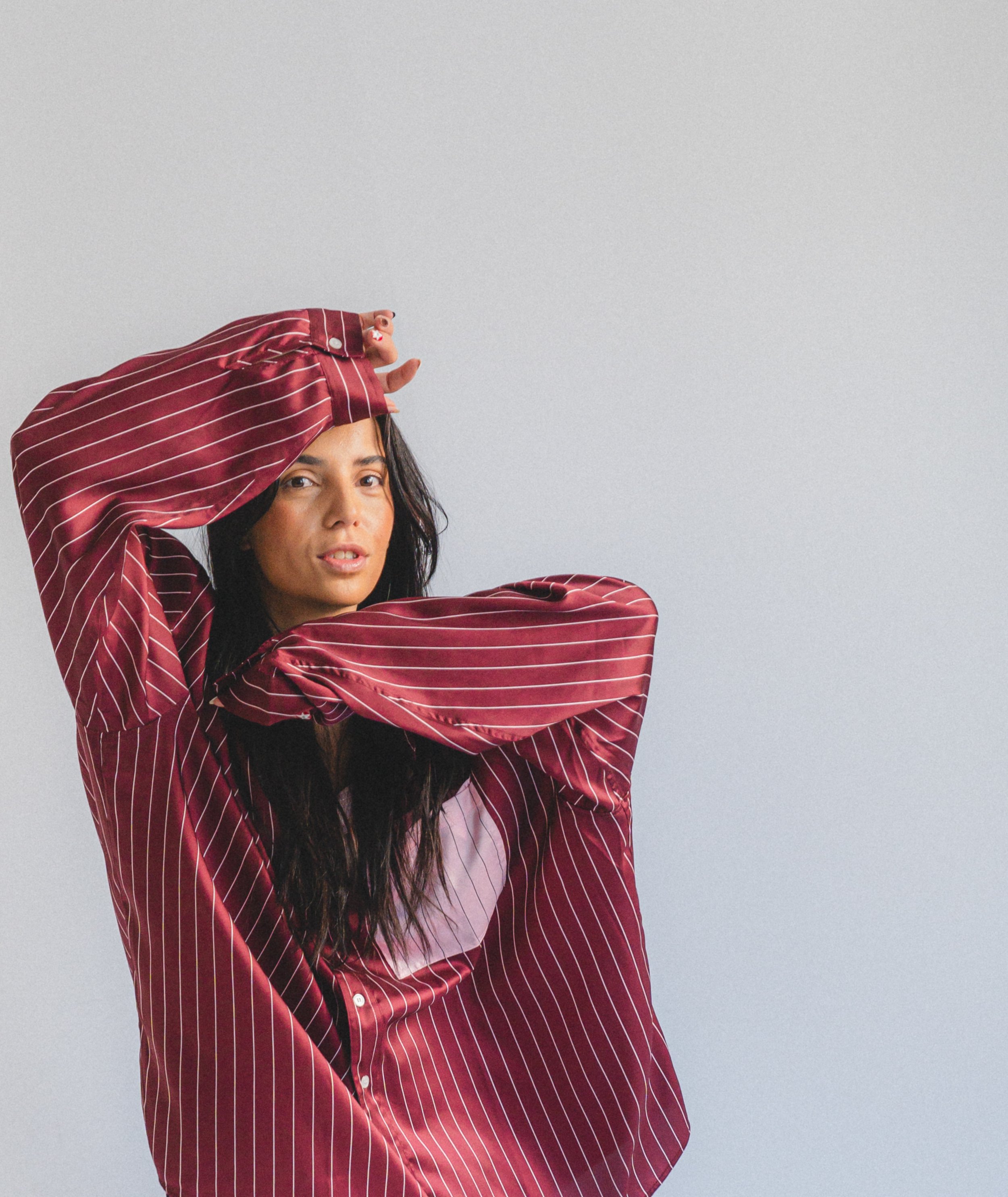 A woman with long dark hair and light skin is wearing a red and white striped long-sleeve shirt. She has one arm across her chest and the other raised with her hand near her forehead, looking slightly upward with a neutral expression. The background 