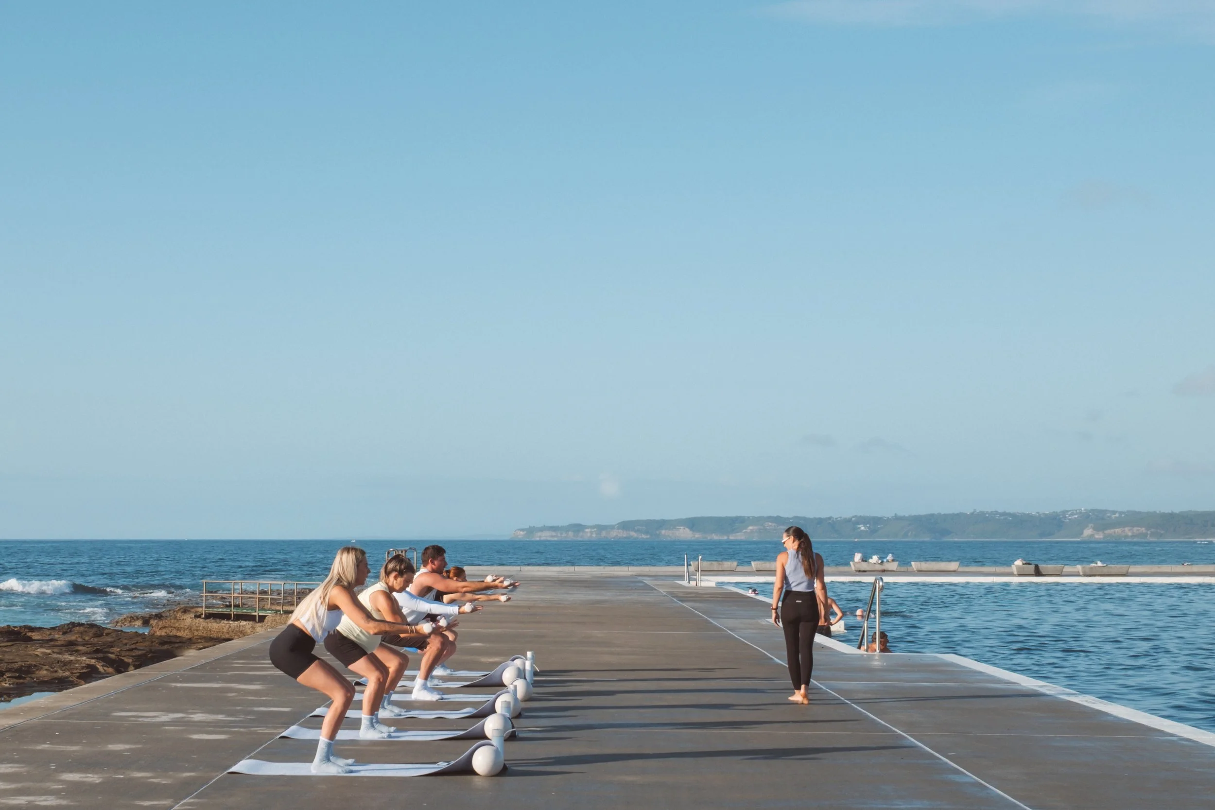 People doing yoga or stretching exercises on a dock by the ocean on a sunny day with clear blue sky and distant shoreline.