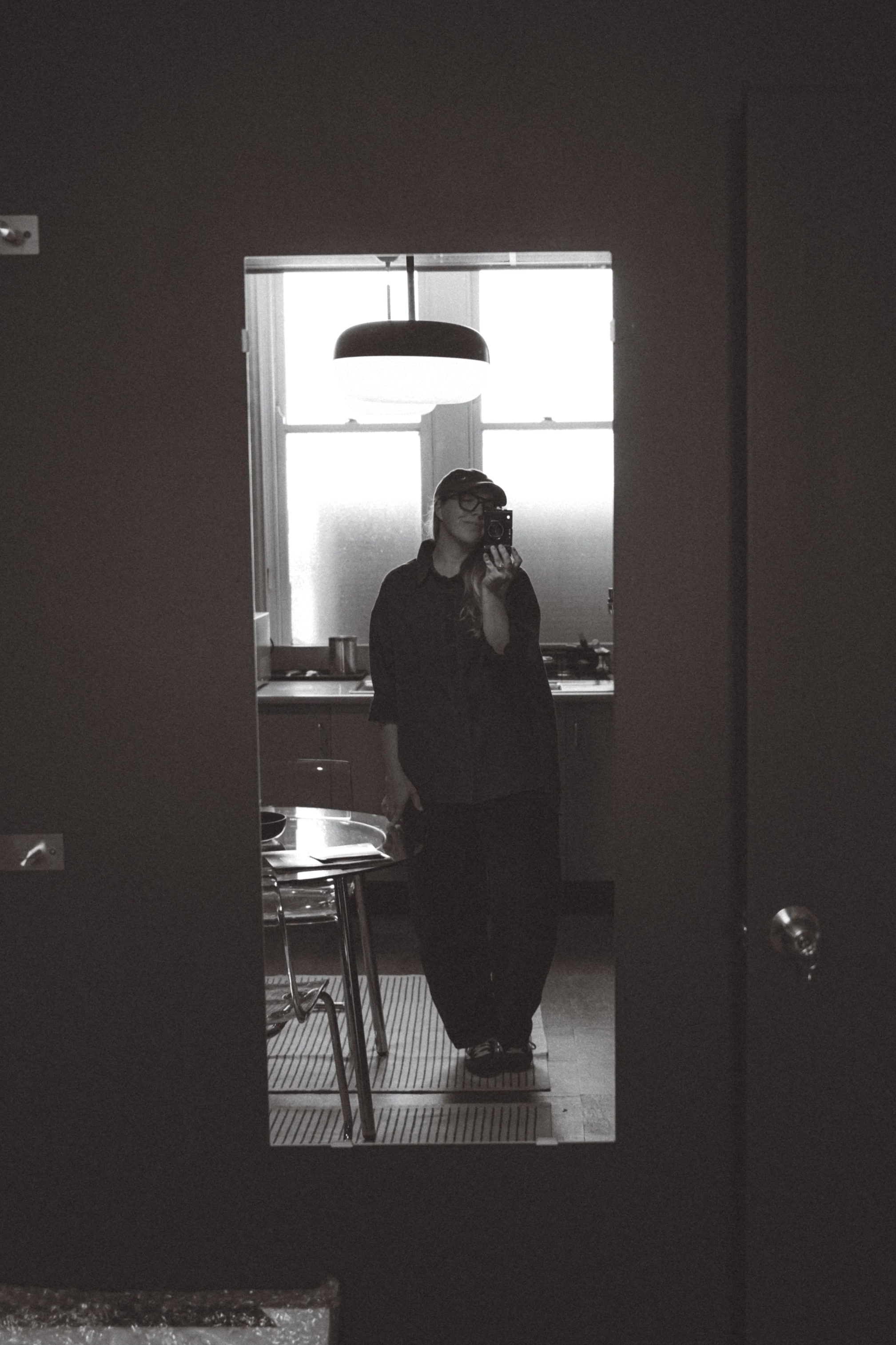 A woman is taking a selfie in a mirror, standing in a kitchen area with a window behind her. She is wearing glasses, a hat, and a dark outfit, and her reflection shows kitchen cabinets and a hanging light fixture.