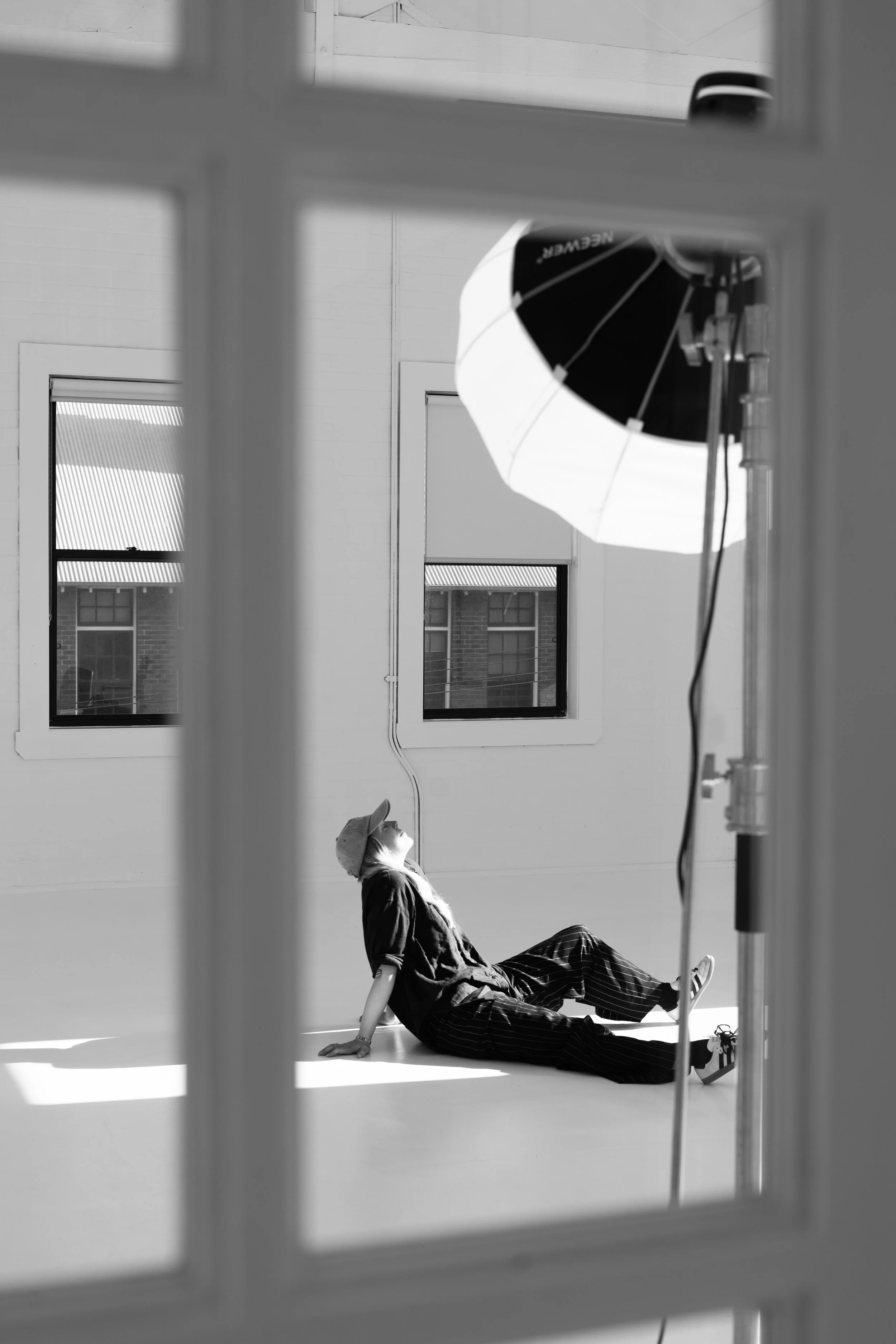A woman in casual clothing and a hat sitting on the floor, leaning back on her hands, inside a photography studio with a large umbrella light.