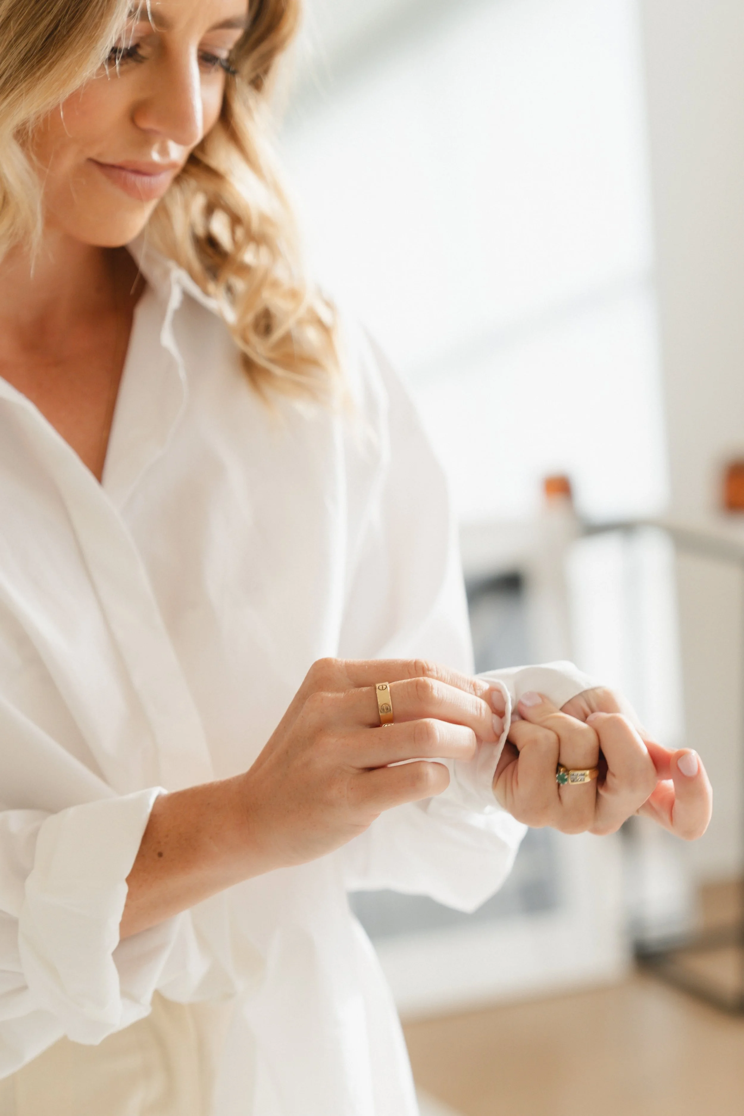 A woman with blonde, curly hair, wearing a white shirt, is buttoning or unbuttoning a shirt cuff. She is wearing multiple rings, including a gold band and a turquoise ring. The background is softly blurred.