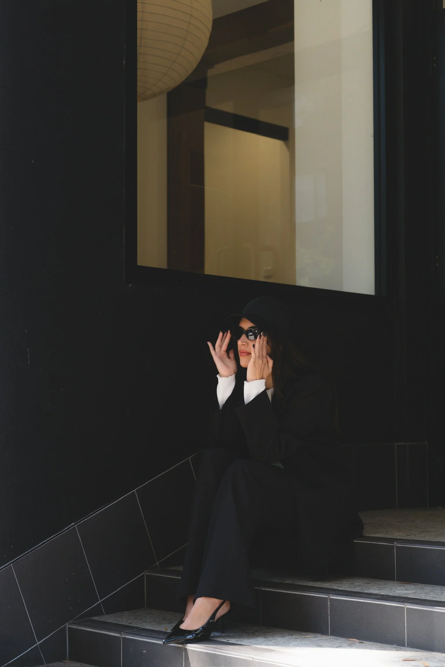 A woman in black attire and heels sitting on stairs, adjusting her black sunglasses, with a large black wall behind her and a window reflecting light.