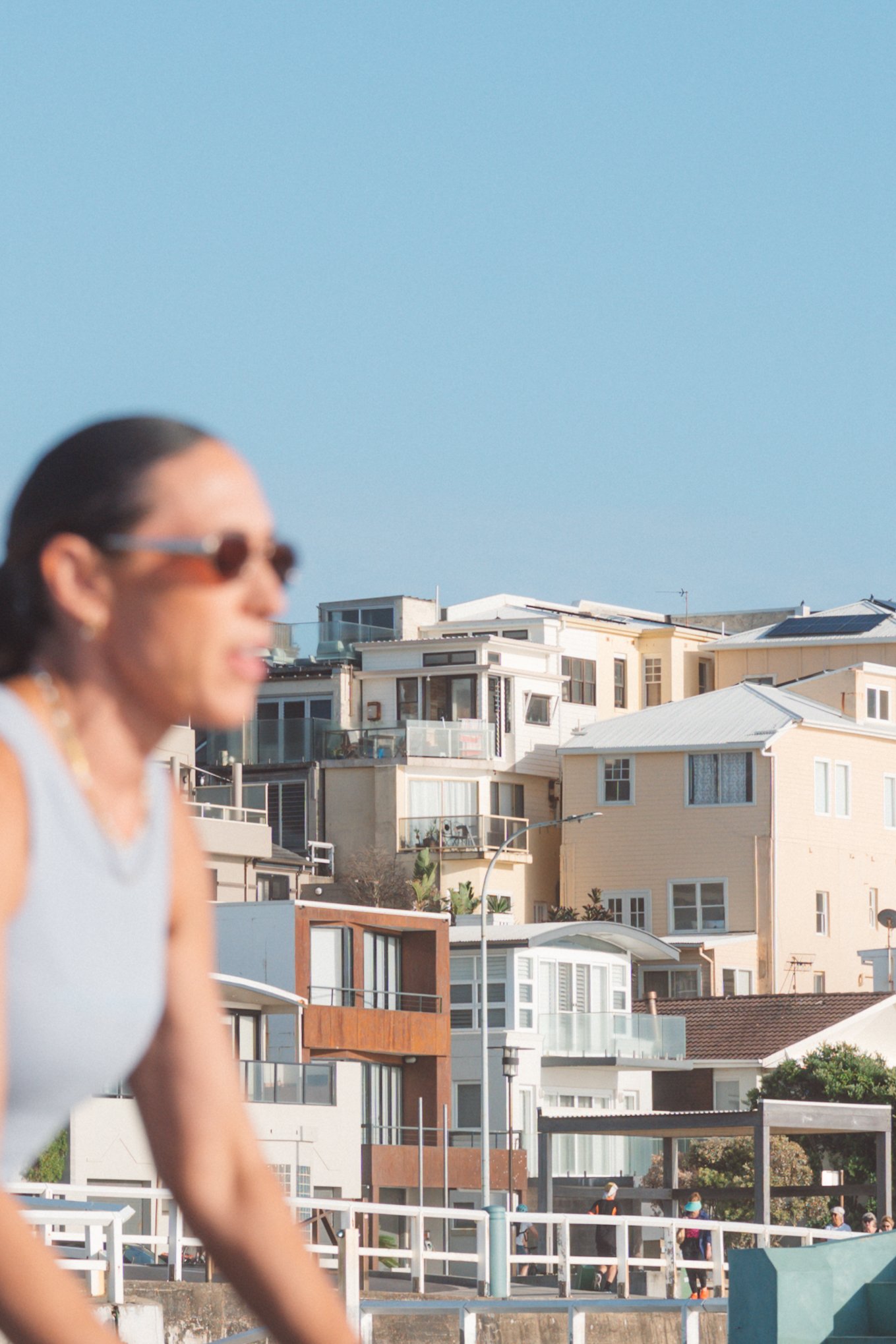 Out-of-focus woman wearing sunglasses in foreground with modern beach houses and a pathway with people walking in the background under a clear blue sky.