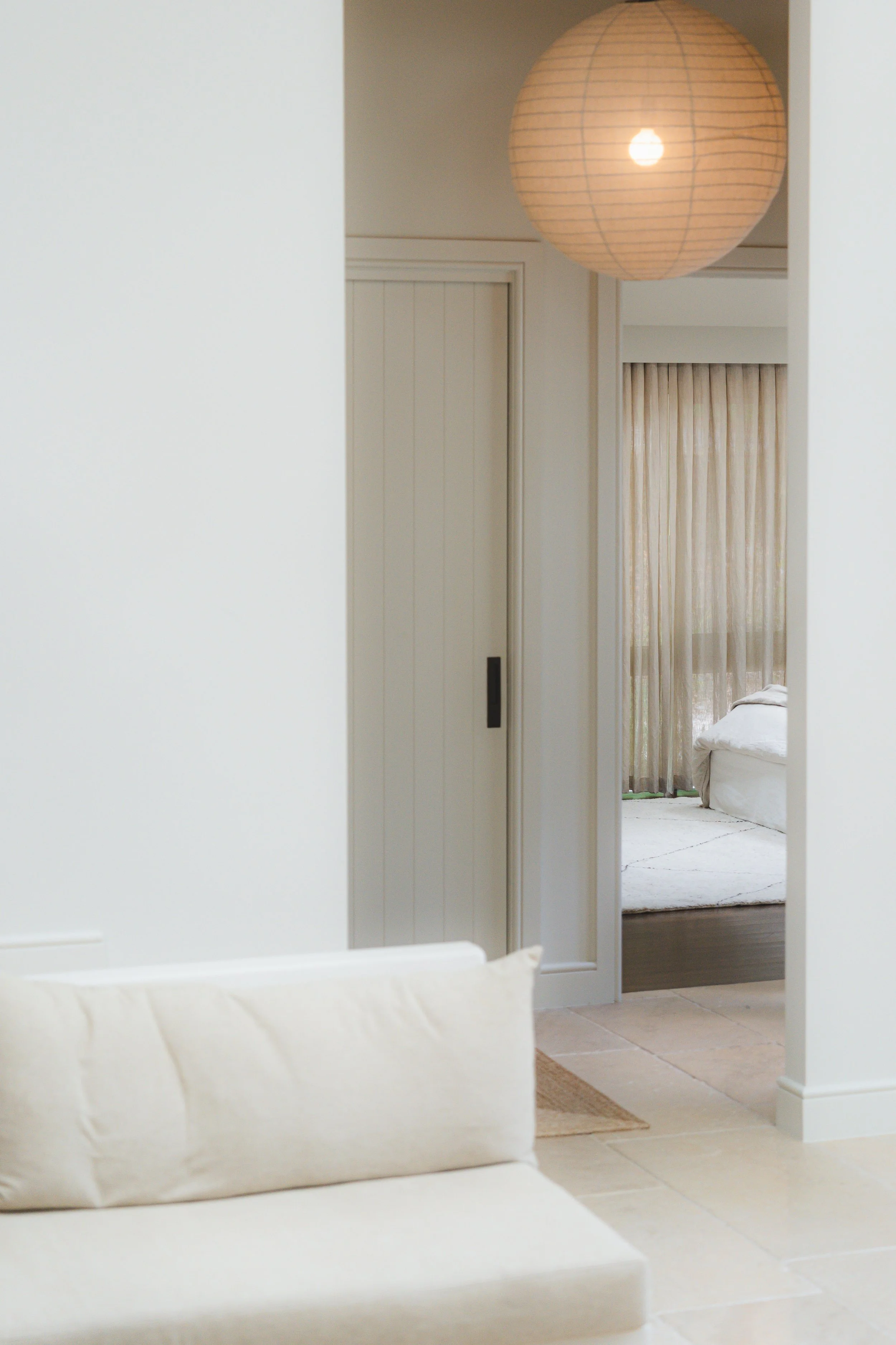 A living room with white walls, a beige tiled floor, and a white couch partially visible in the foreground. Through an open doorway, a bedroom with a bed covered in white bedding, beige/cream curtains, and a round paper lantern hanging from the ceili