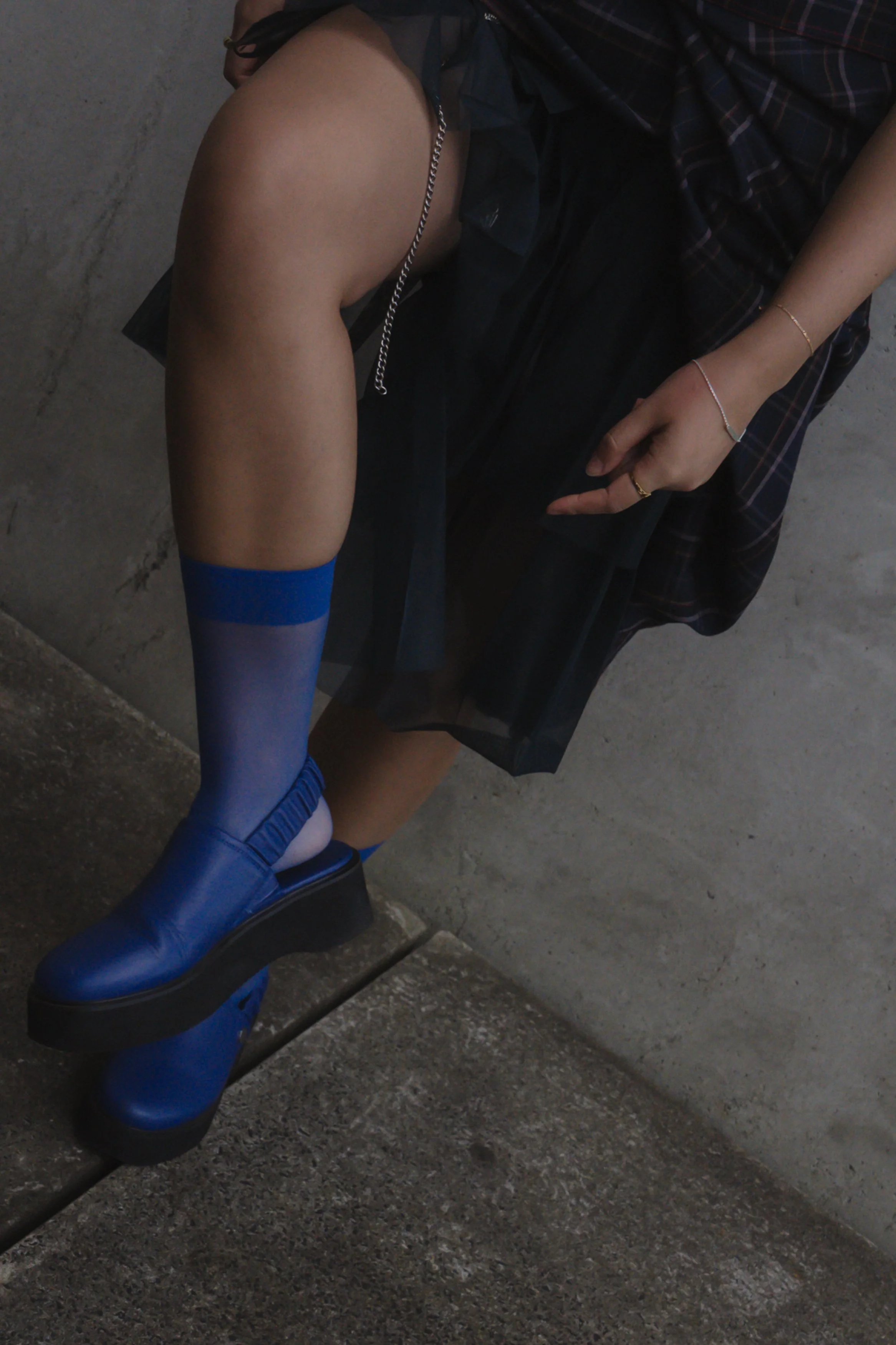 Close-up of a person's legs wearing blue platform boots, a plaid skirt, and wire bracelets, with part of their hand resting on their thigh, against a concrete background.