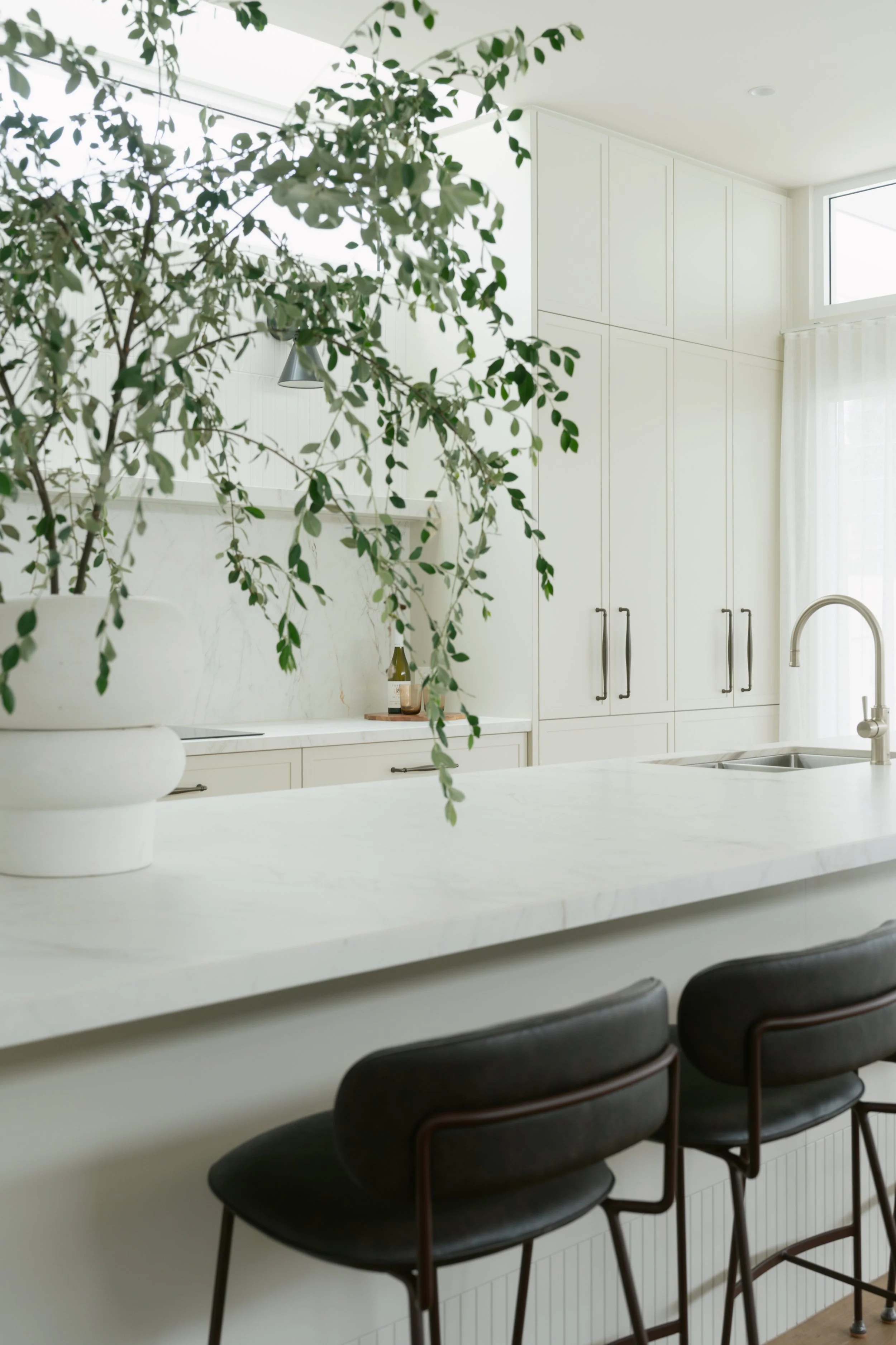 A modern kitchen with white cabinetry, a white marble countertop, and black barstools with dark wooden frames lined up along a white breakfast bar. There is a potted plant on the counter and natural light coming through a window with a curtain.