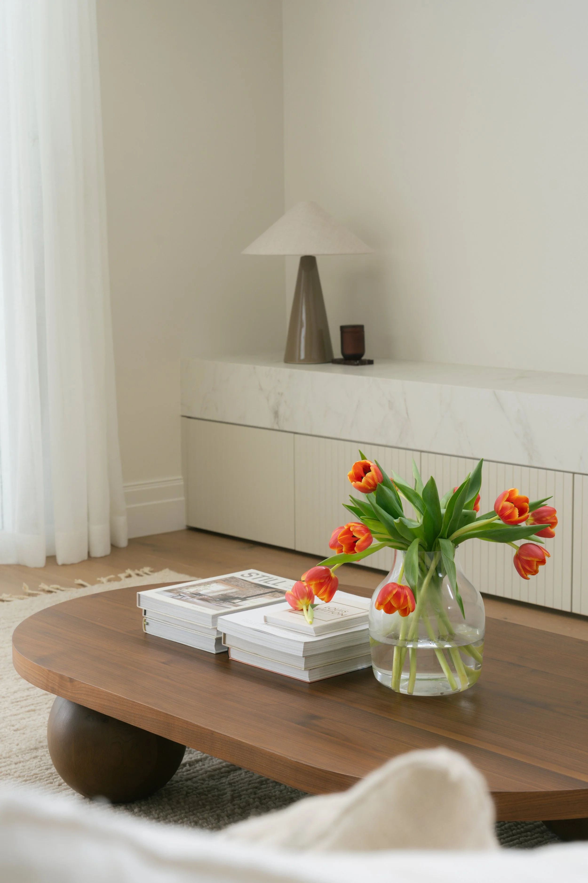 A wooden coffee table with a glass vase of orange tulips and several magazines or books on it, in a modern living room with a white marble sideboard and a lamp in the background.