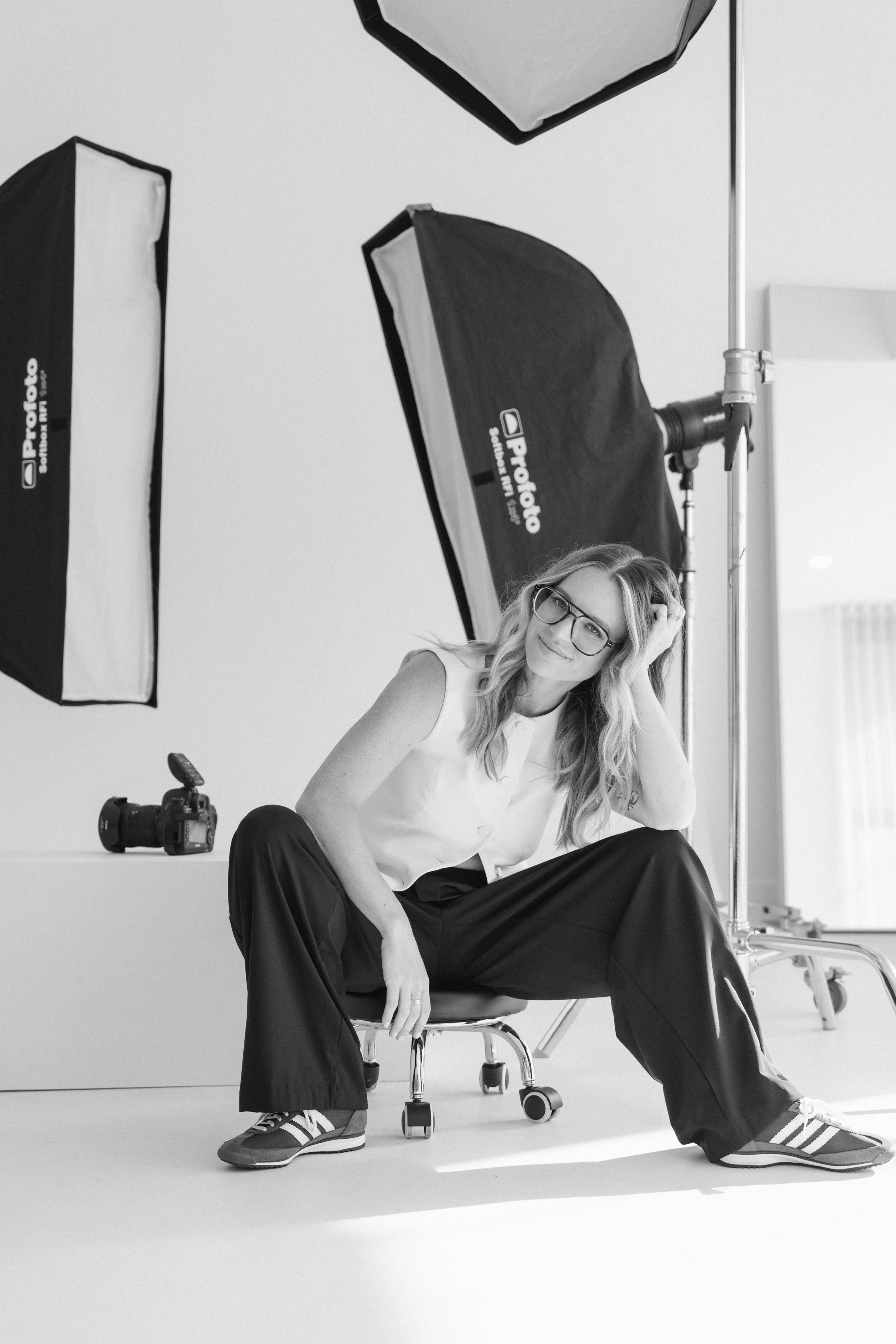 A woman with glasses and long wavy hair, dressed in a sleeveless top and wide-legged pants, sitting on a white chair in a photography studio. Around her are professional lighting equipment, including softbox lights, and a camera on the floor, with a 