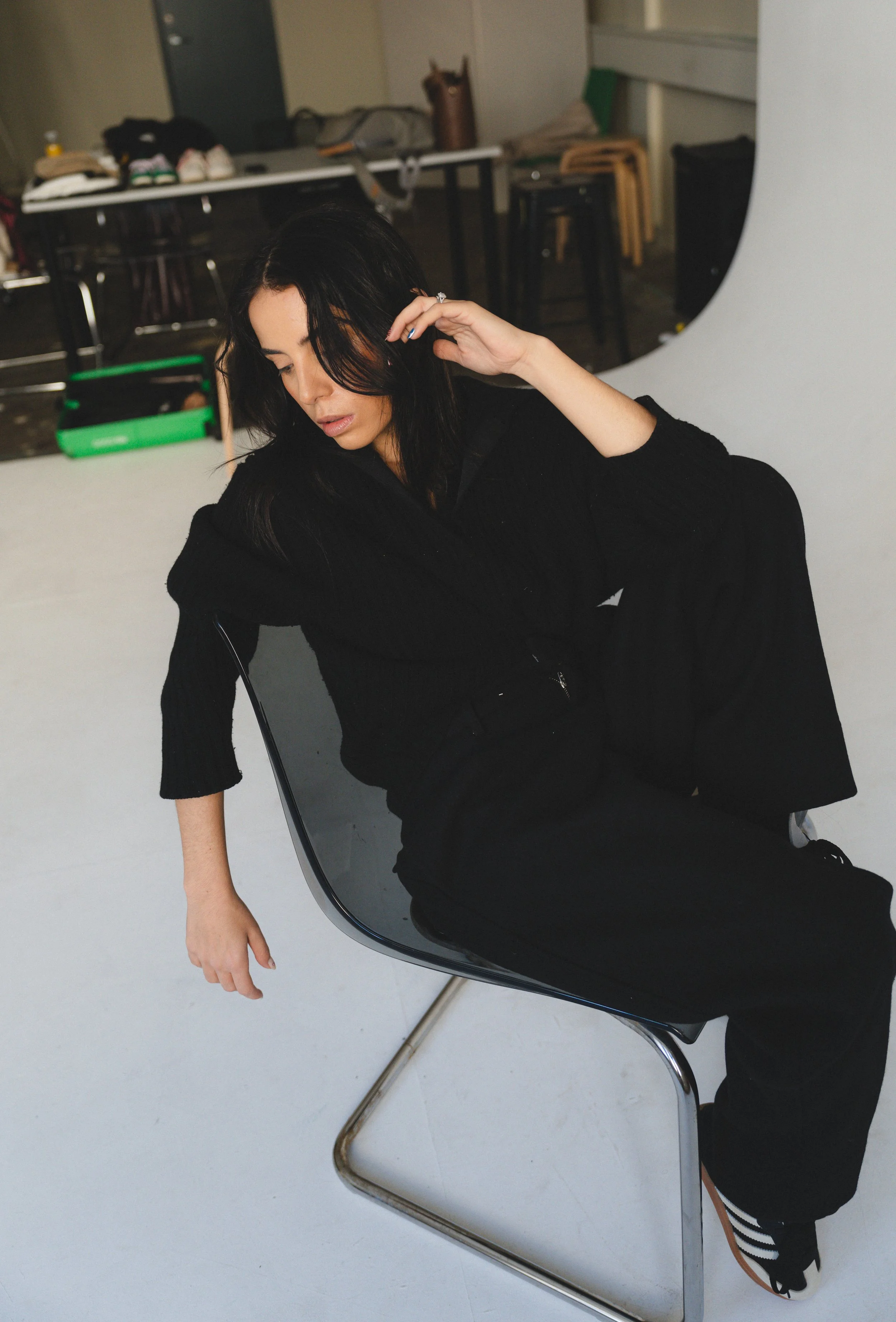 A woman with black hair, dressed in black, is sitting in a modern black chair with a metallic frame, in a photography studio with a white backdrop. She is resting her arm on her knee, with a thoughtful expression, and appears to be adjusting her hair