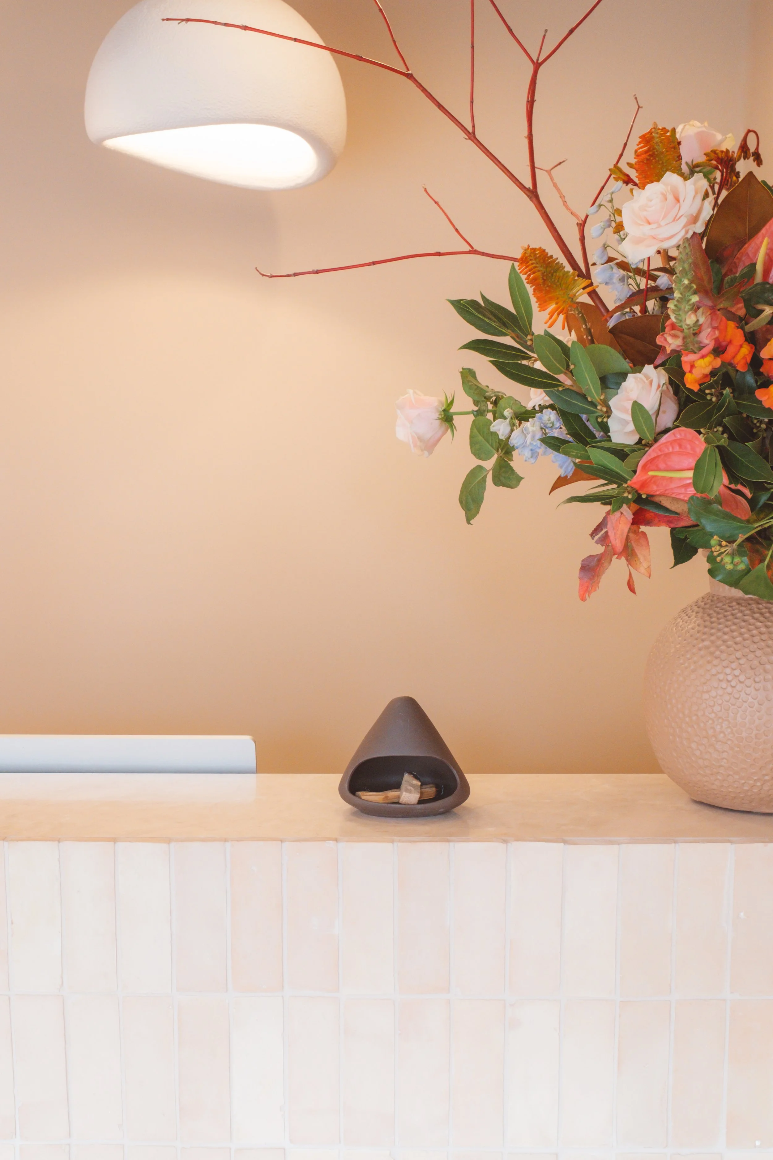 A beige wall with a modern white pendant light hanging above, a large beige textured vase with a floral arrangement, a small black incense holder with incense sticks and stones, all placed on a light-colored tiled surface.