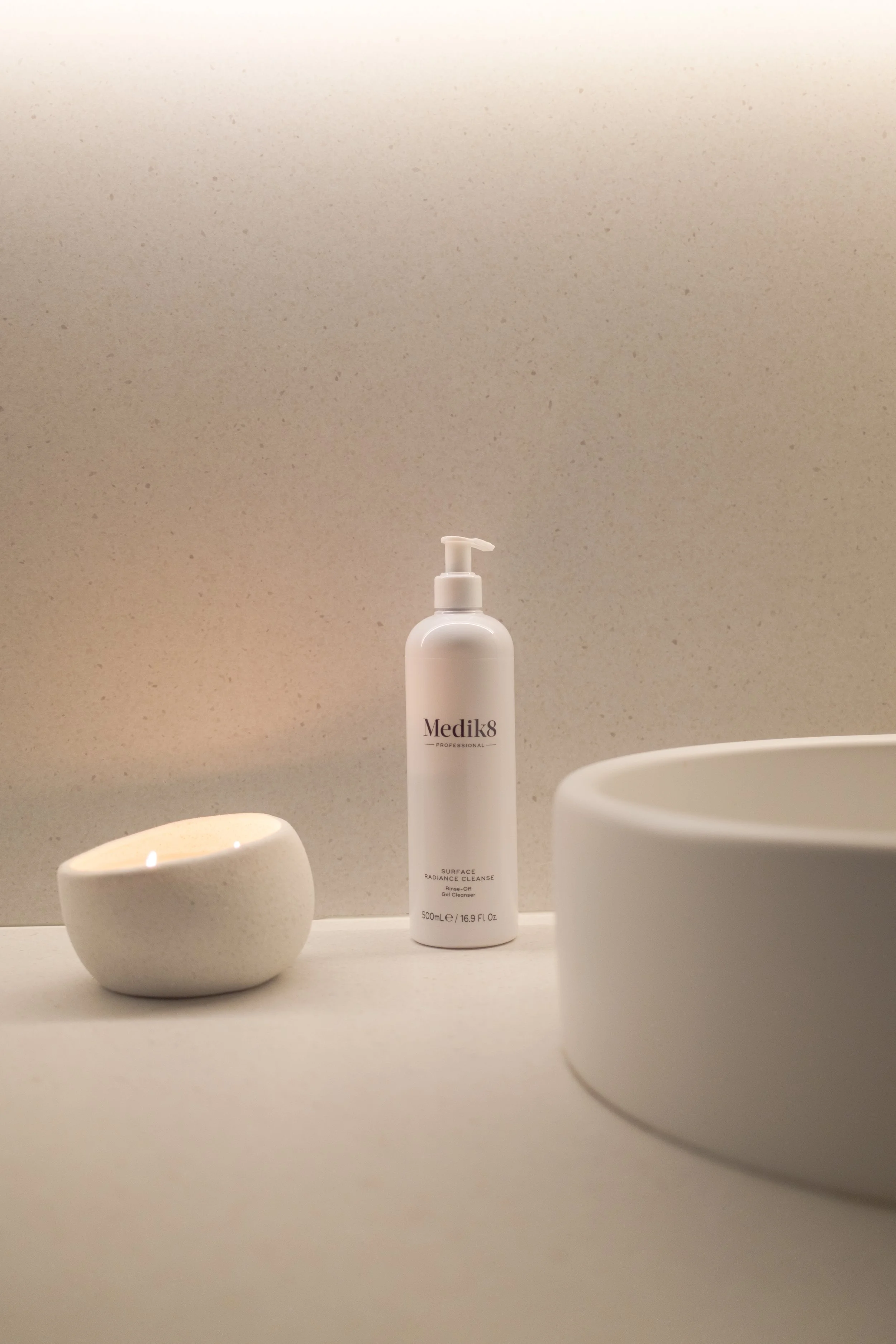 A white bottle of Medik8 Surface Radiance Cleanse on a white surface, surrounded by white bowls, set against a beige speckled background.