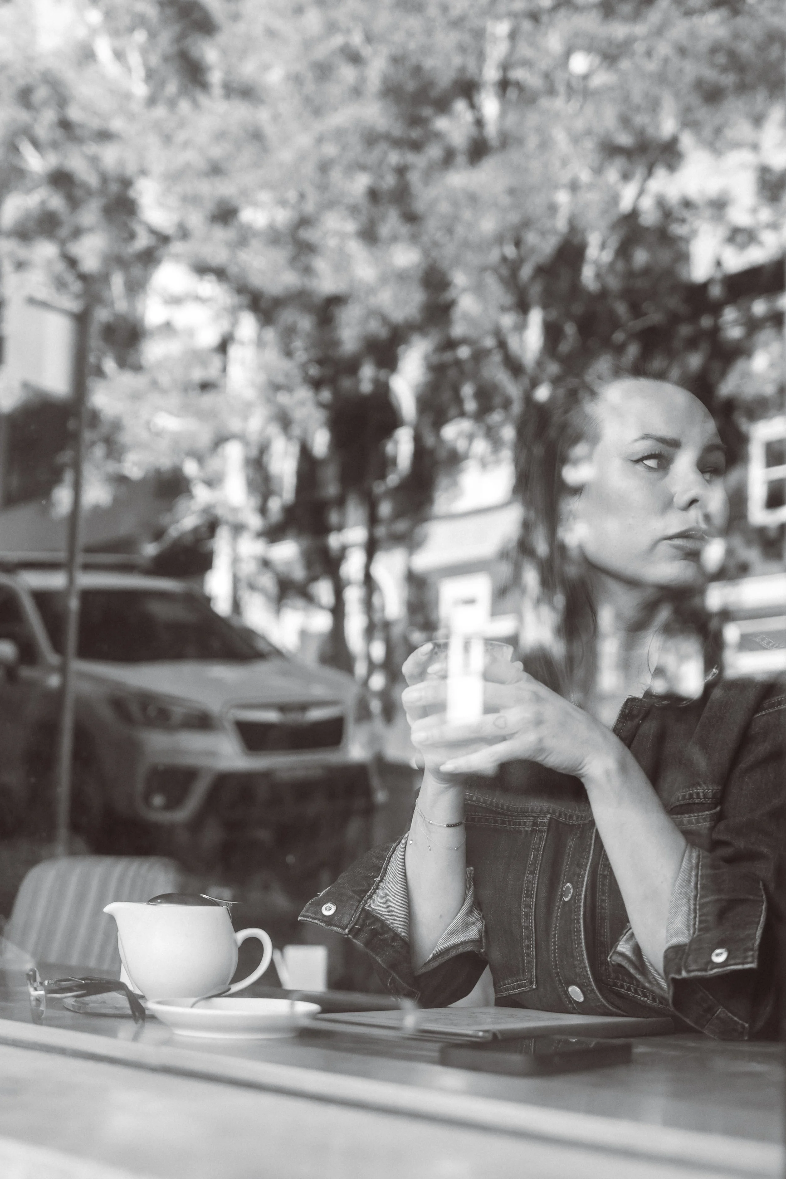 A woman with dark hair and a denim jacket sitting at a table, holding a glass with a drink. There is a white teacup and saucer on the table, and a pair of sunglasses. Outside, through the window, there are cars parked on the street and blurred trees 