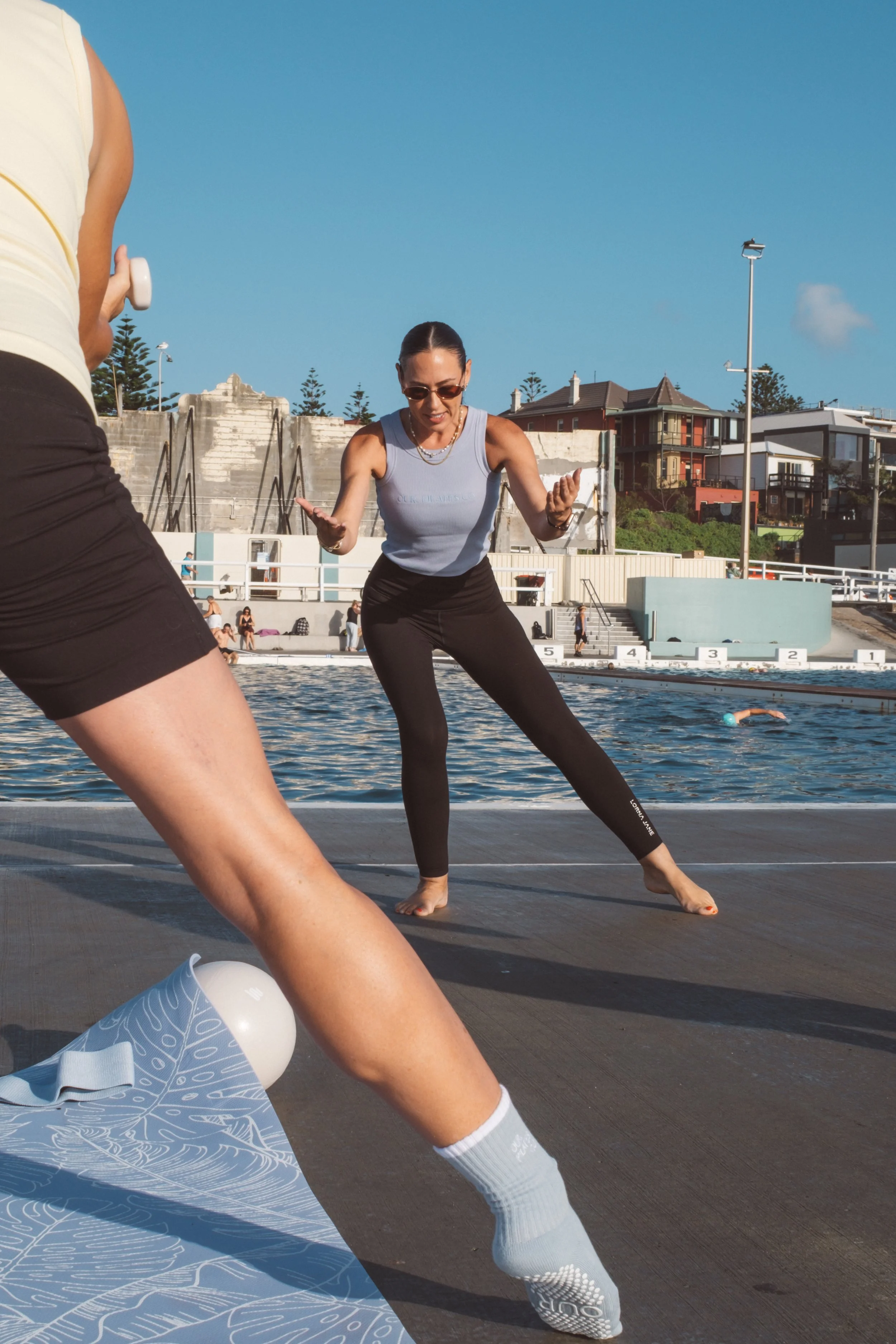 A woman practicing yoga outdoors by a swimming pool with a scenic hillside background, wearing sunglasses, a gray sleeveless top, and black leggings.