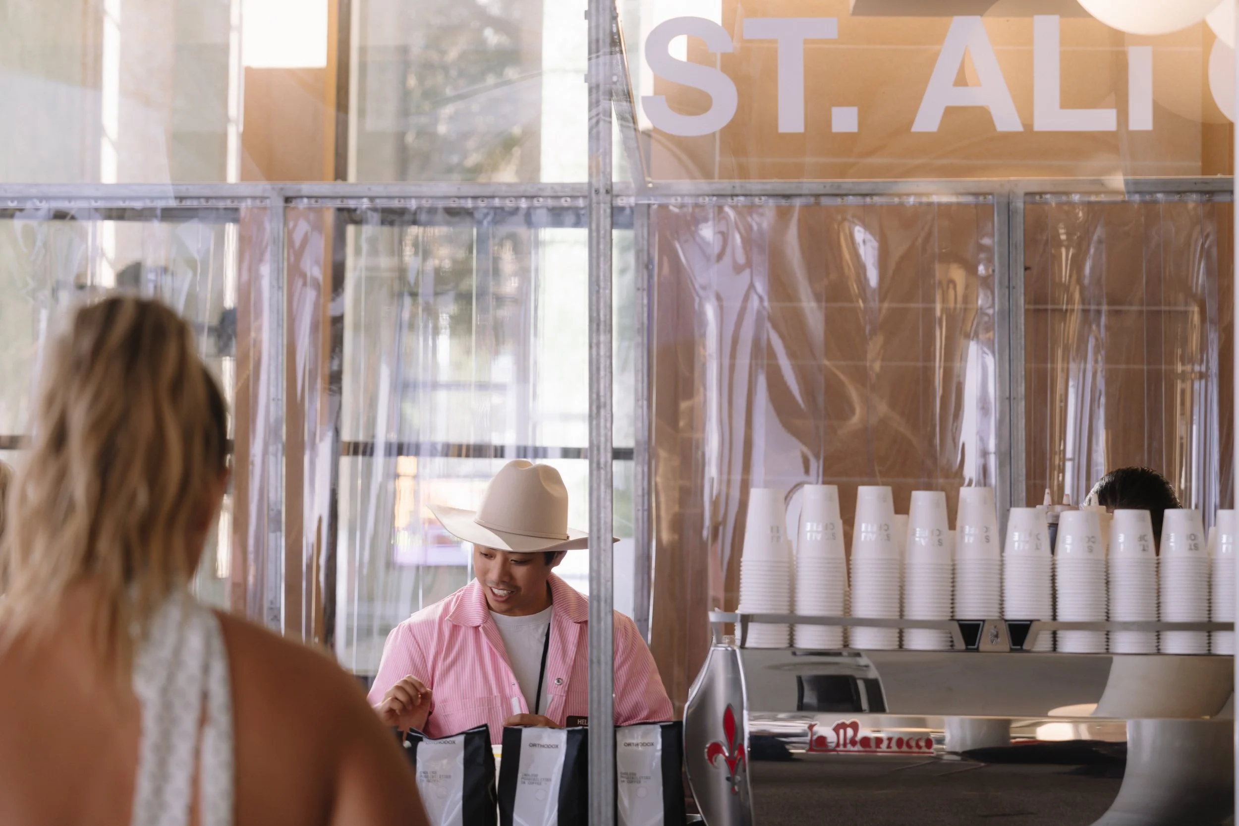 A woman with blonde hair is facing a counter where a smiling man wearing a pink shirt and cowboy hat is serving her. The background shows stacks of white cups and a sign with 'ST. AL' partially visible.