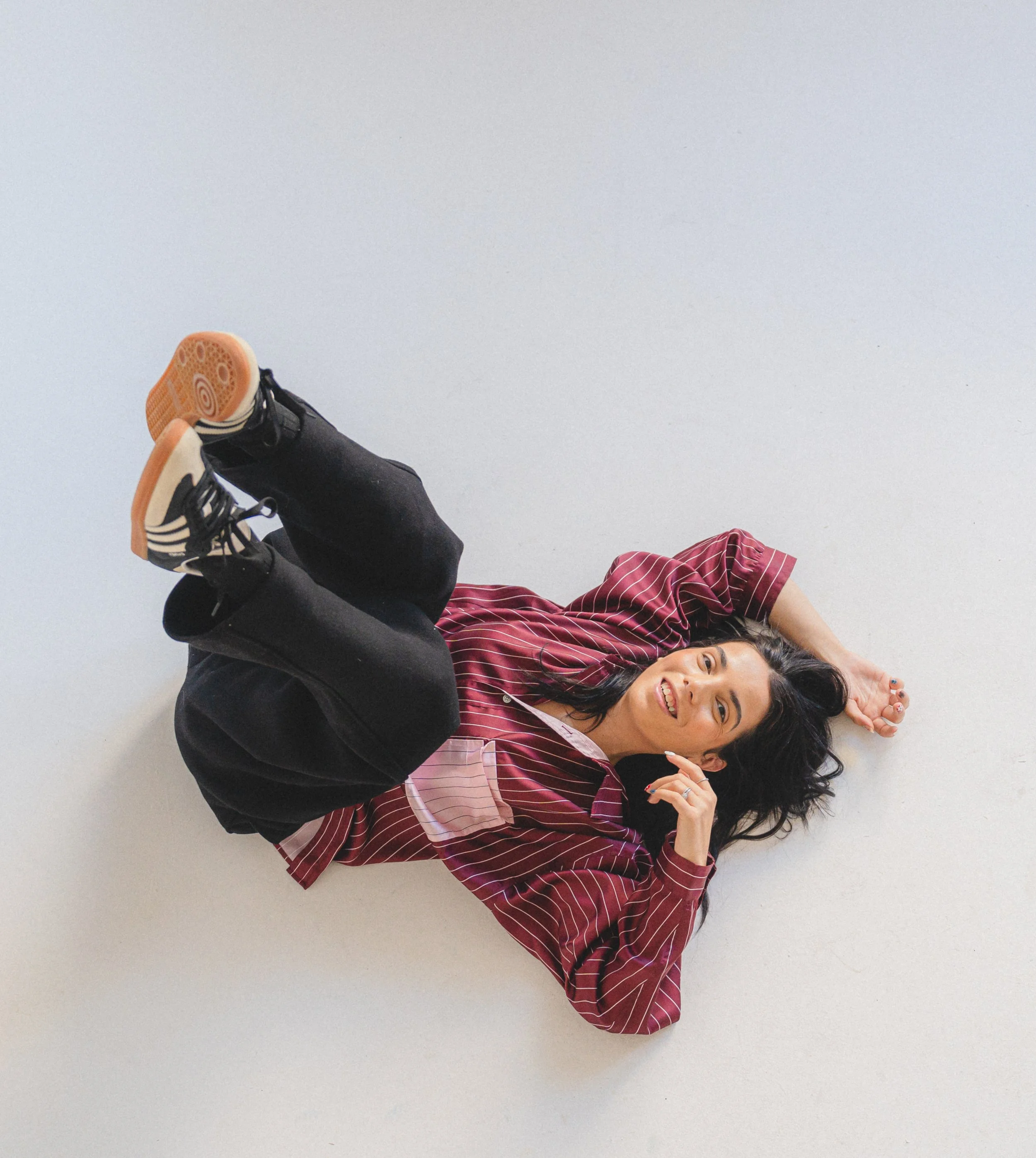 A person lying on a white floor, smiling and making a peace sign with one hand. They are wearing a red and white striped shirt and black pants. Their shoes are black with a beige sole and black laces.