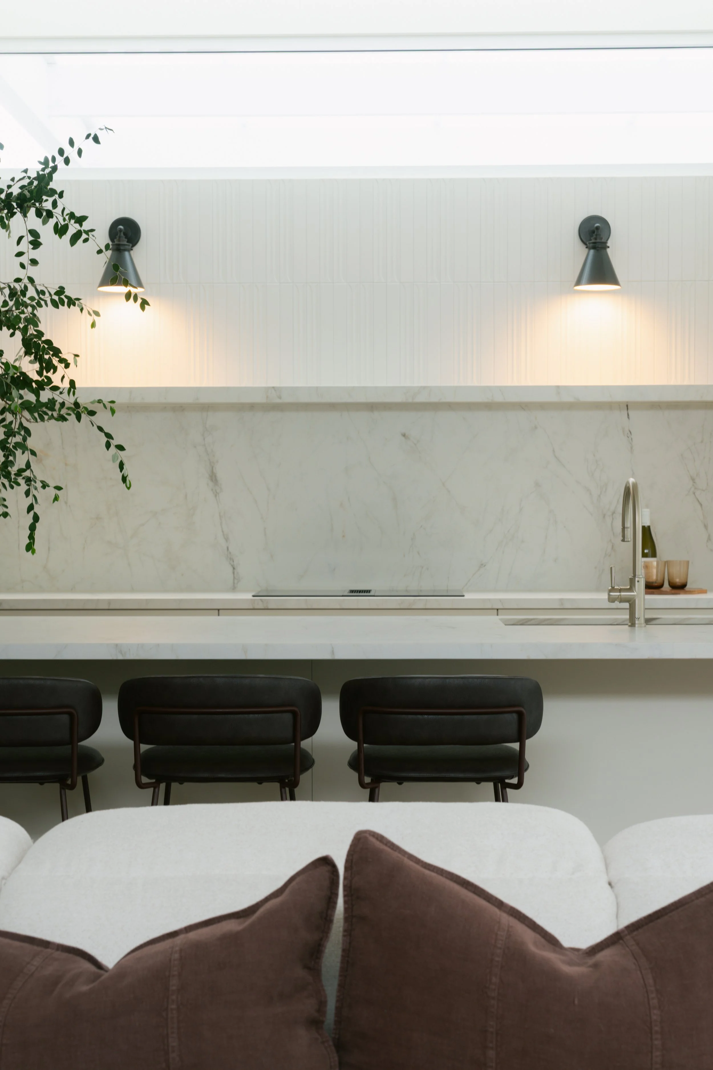 Modern kitchen with white marble countertops, white tiled backsplash, black dining chairs, and wall-mounted black lamps