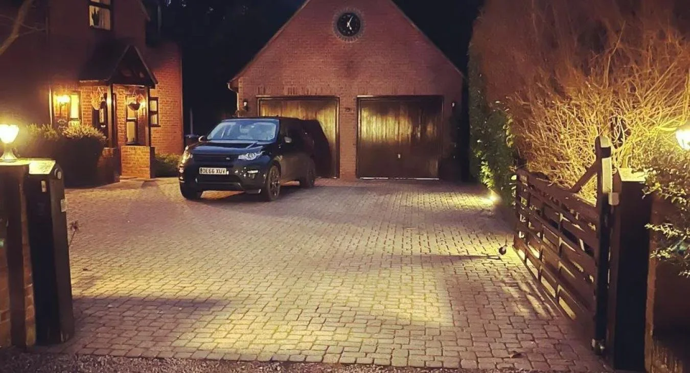 A driveway with a black car parked in front of a garage on a brick house at night, illuminated by security outdoor lights.