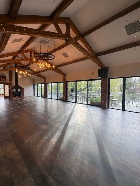 The interior of Willow Barn at Bennetts Farm, Worcester — a spacious barn with exposed wooden beams, warm Edison lighting and large glass doors overlooking greenery, home to Yin & Tonic with Women & Wild.