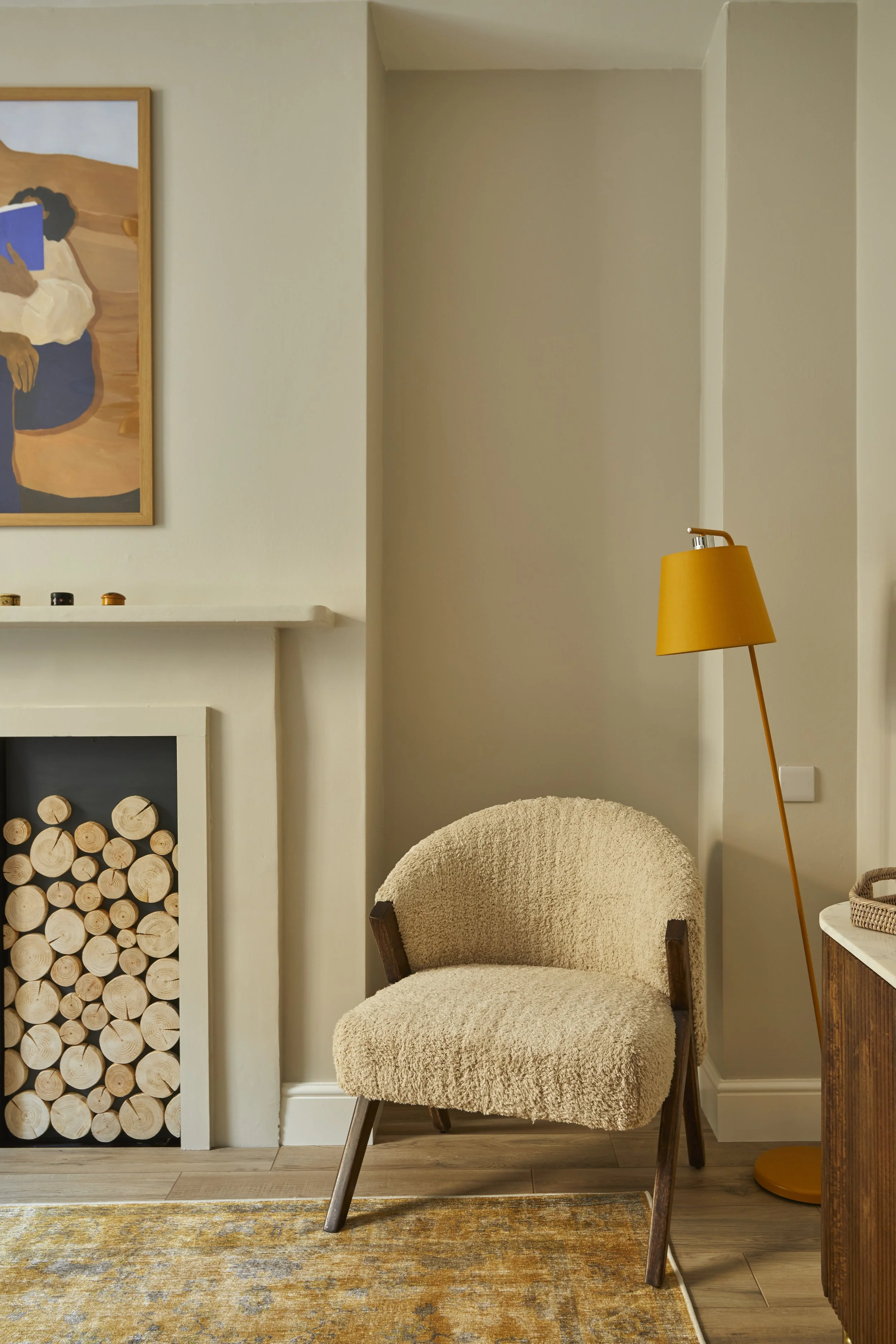 A cozy corner with a plush beige armchair, a mustard-yellow floor lamp, and a fireplace with stacked firewood, alongside a framed artwork on the wall.
