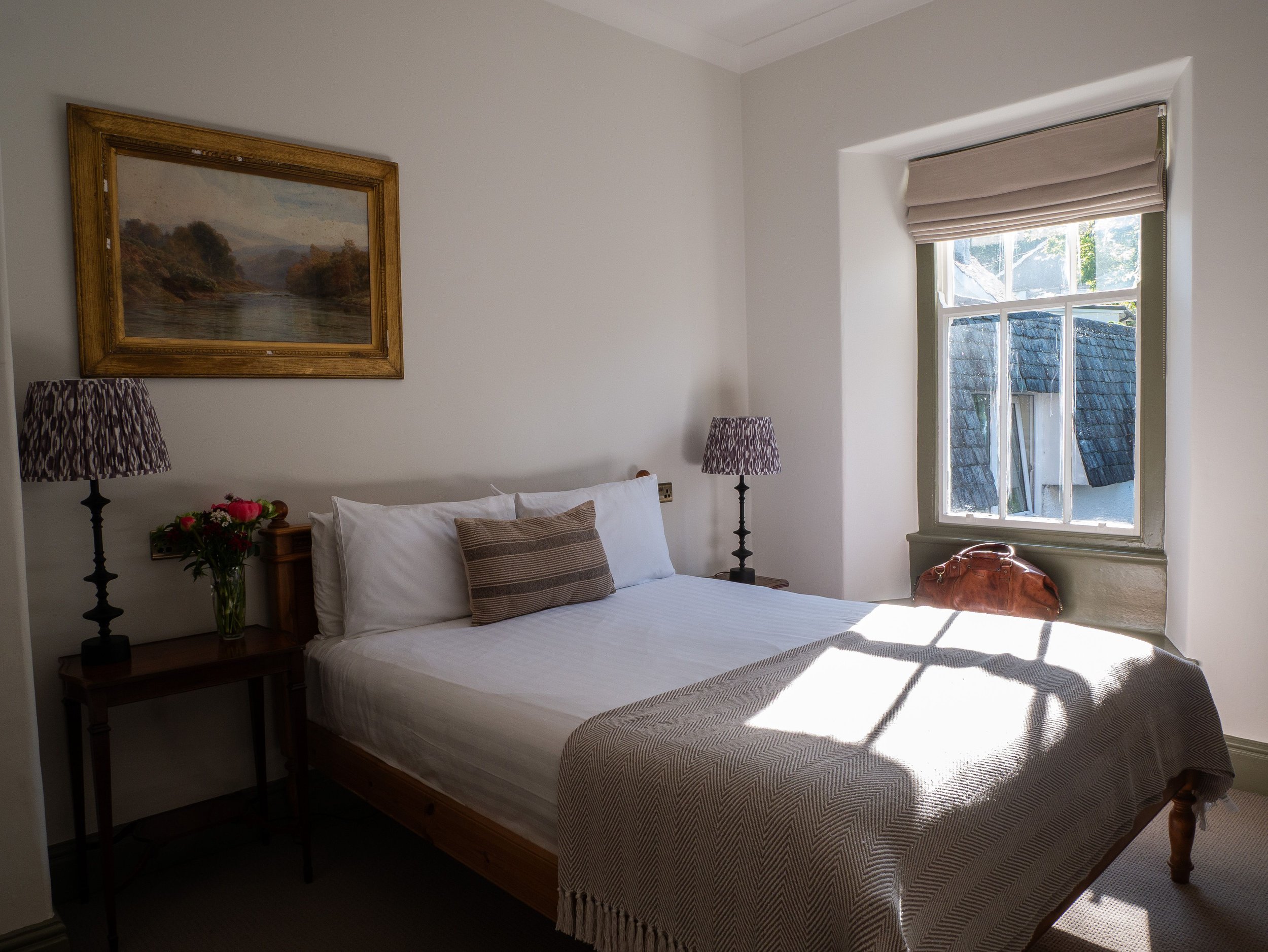 A cozy bedroom with a white bed, a brown striped pillow, and two purple lamps on dark wooden nightstands, a window letting in sunlight, and a landscape painting above the bed.