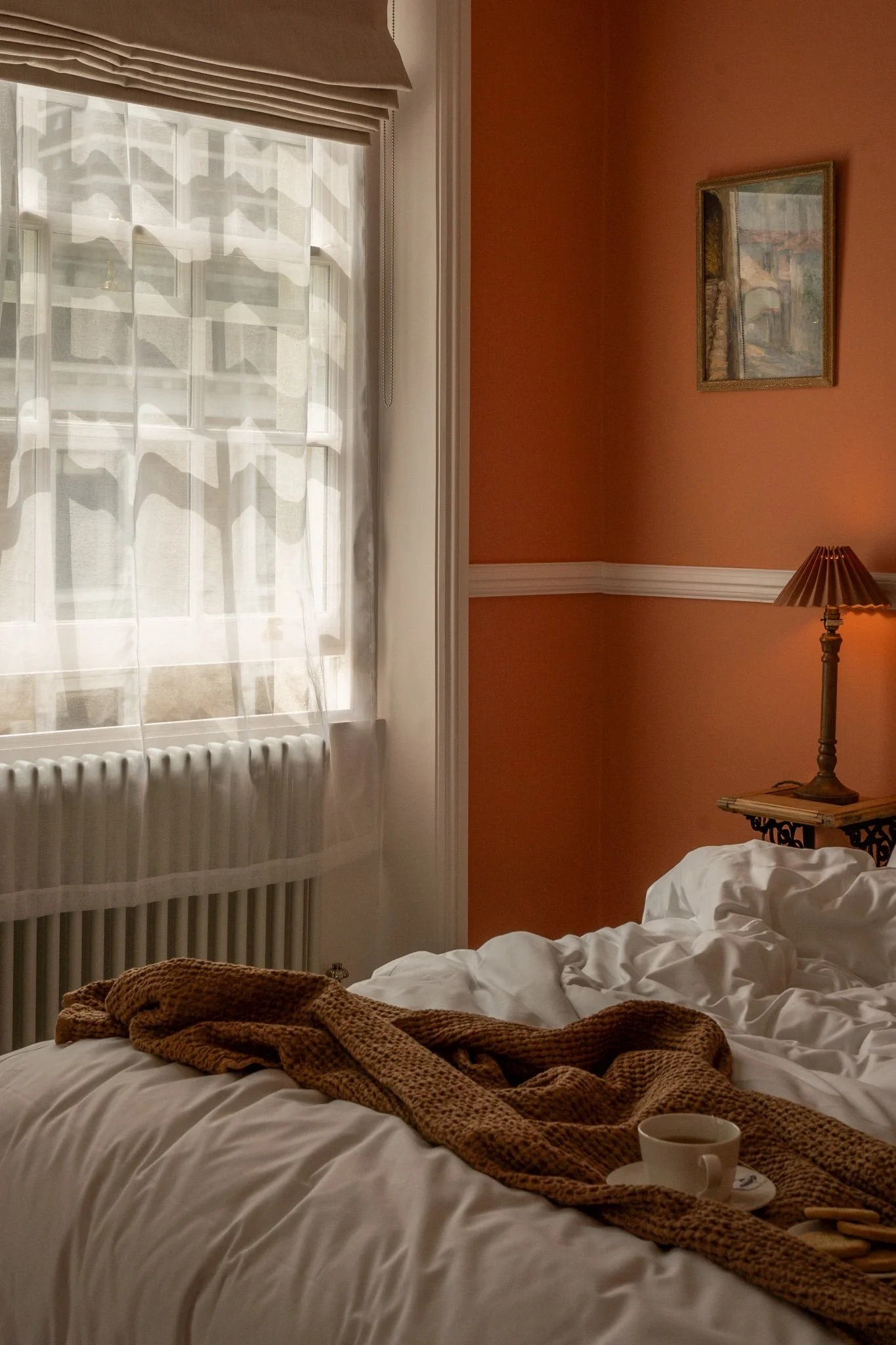 A cozy bedroom scene with sunlight filtering through sheer curtains, a rumpled white bed with a brown knitted blanket, a cup on a tray, and a small lamp on a side table. A framed picture hangs on the orange wall.
