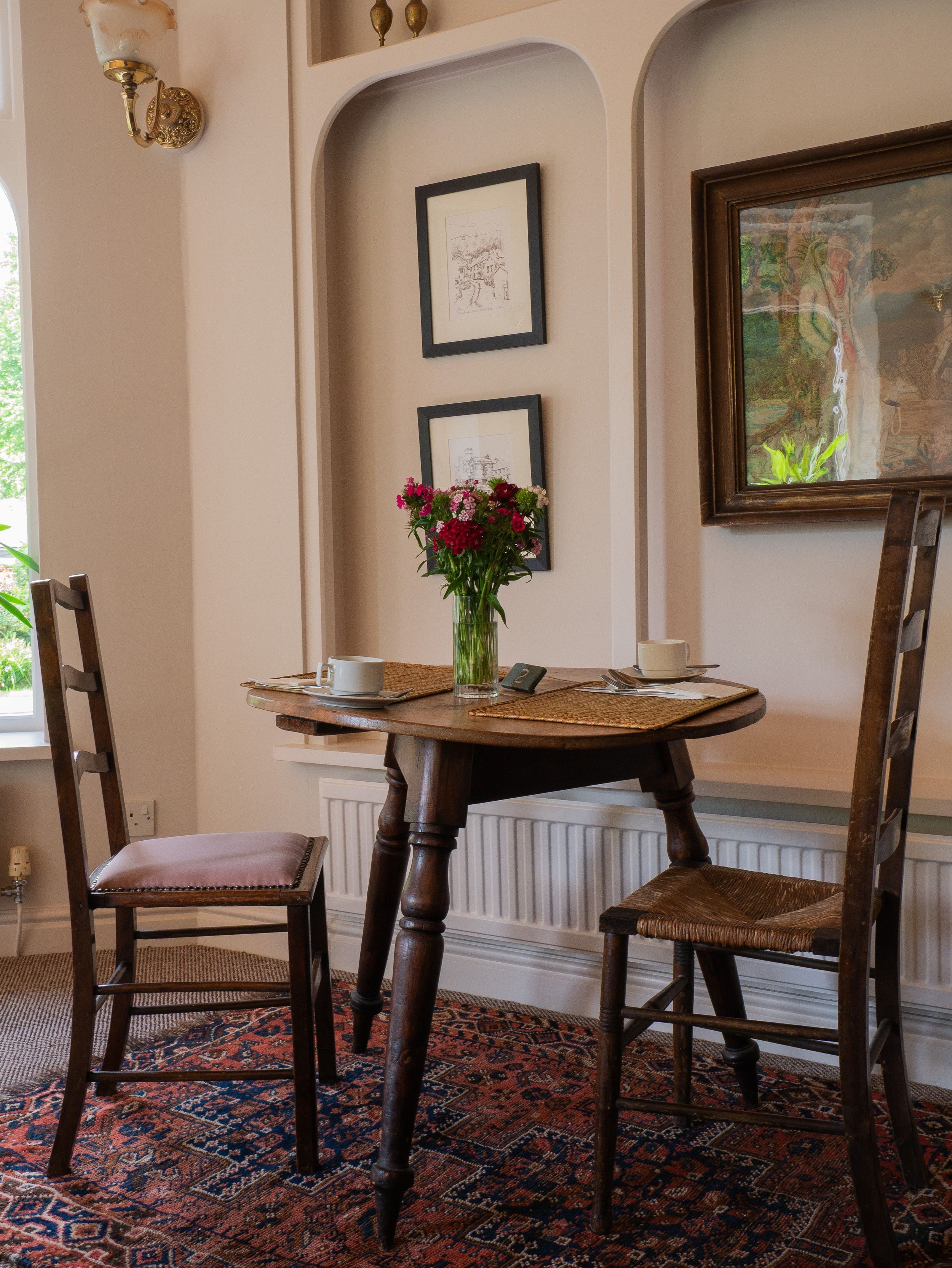 A dining nook with a wooden table set for two, featuring a vase of pink and red flowers, teacups, saucers, and cutlery, with two chairs, framed pictures on the wall, and a patterned rug under the table.