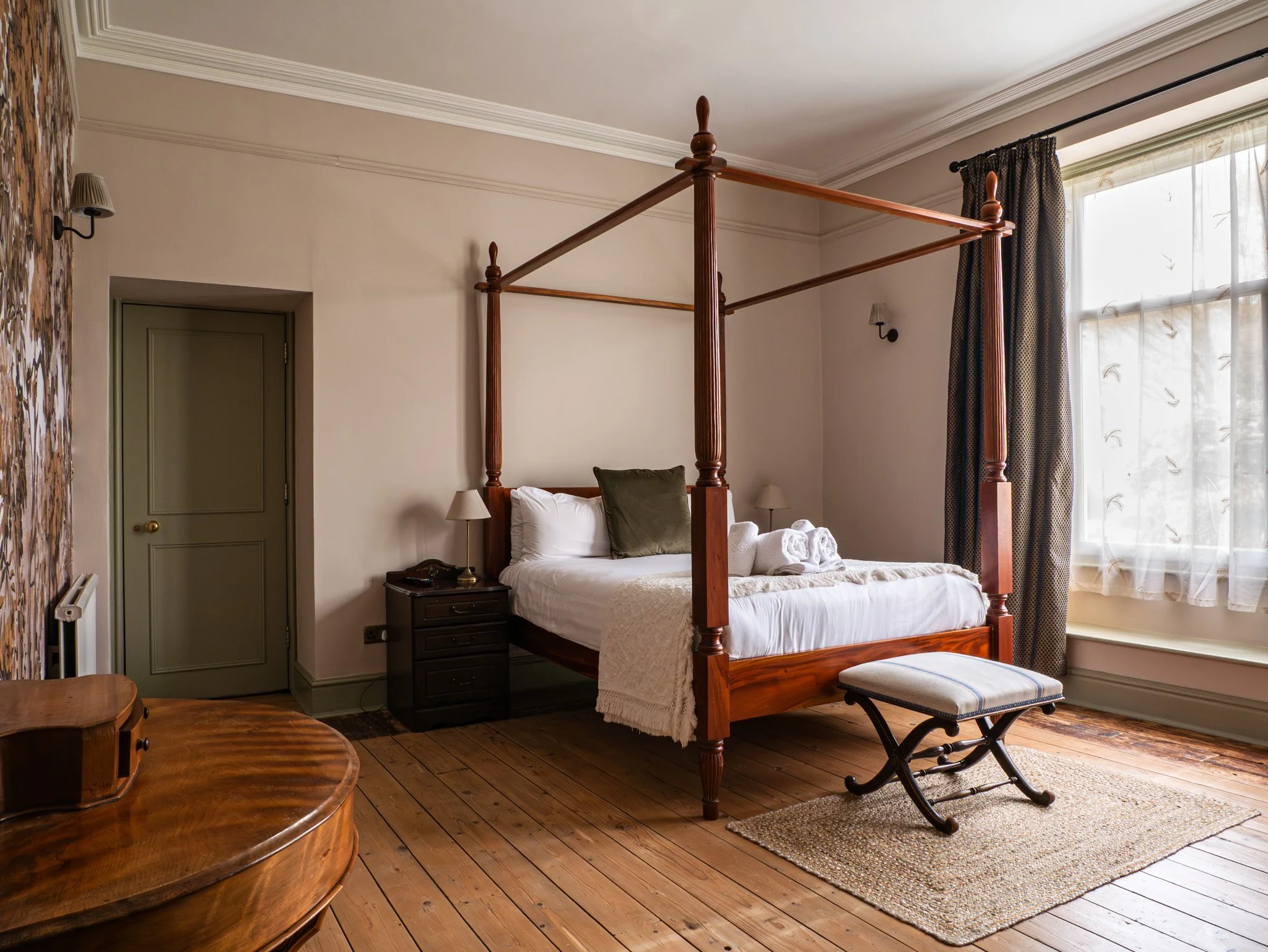 A bedroom with a four-poster wooden bed, a small black bedside table, a bench at the foot of the bed, and a window with curtains letting in natural light.