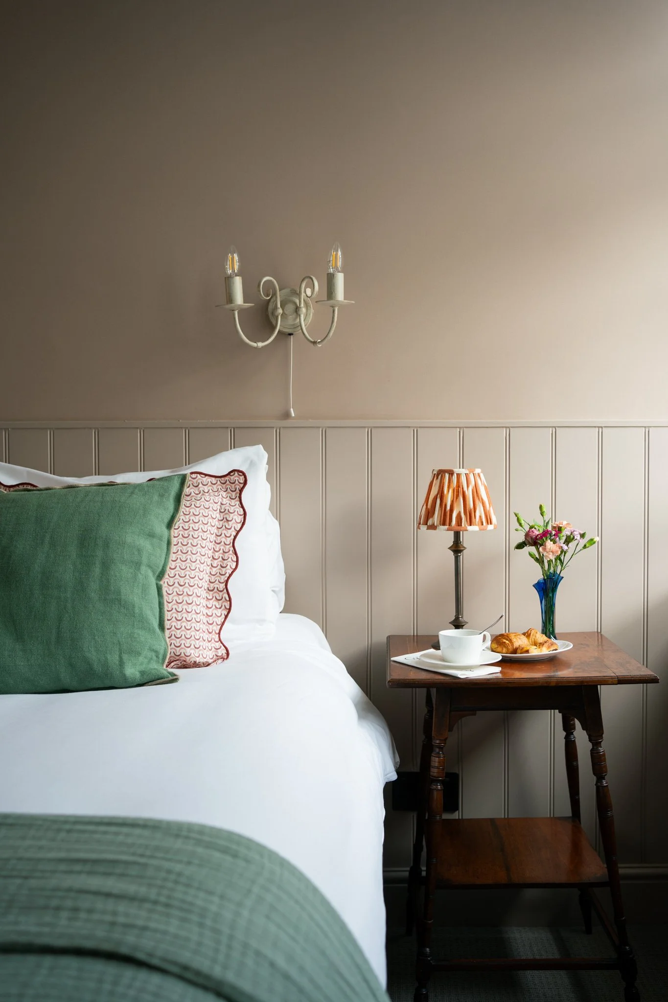 Bedroom corner with bed, pillows, nightstand, lamp, flower vase, cup, and croissants.