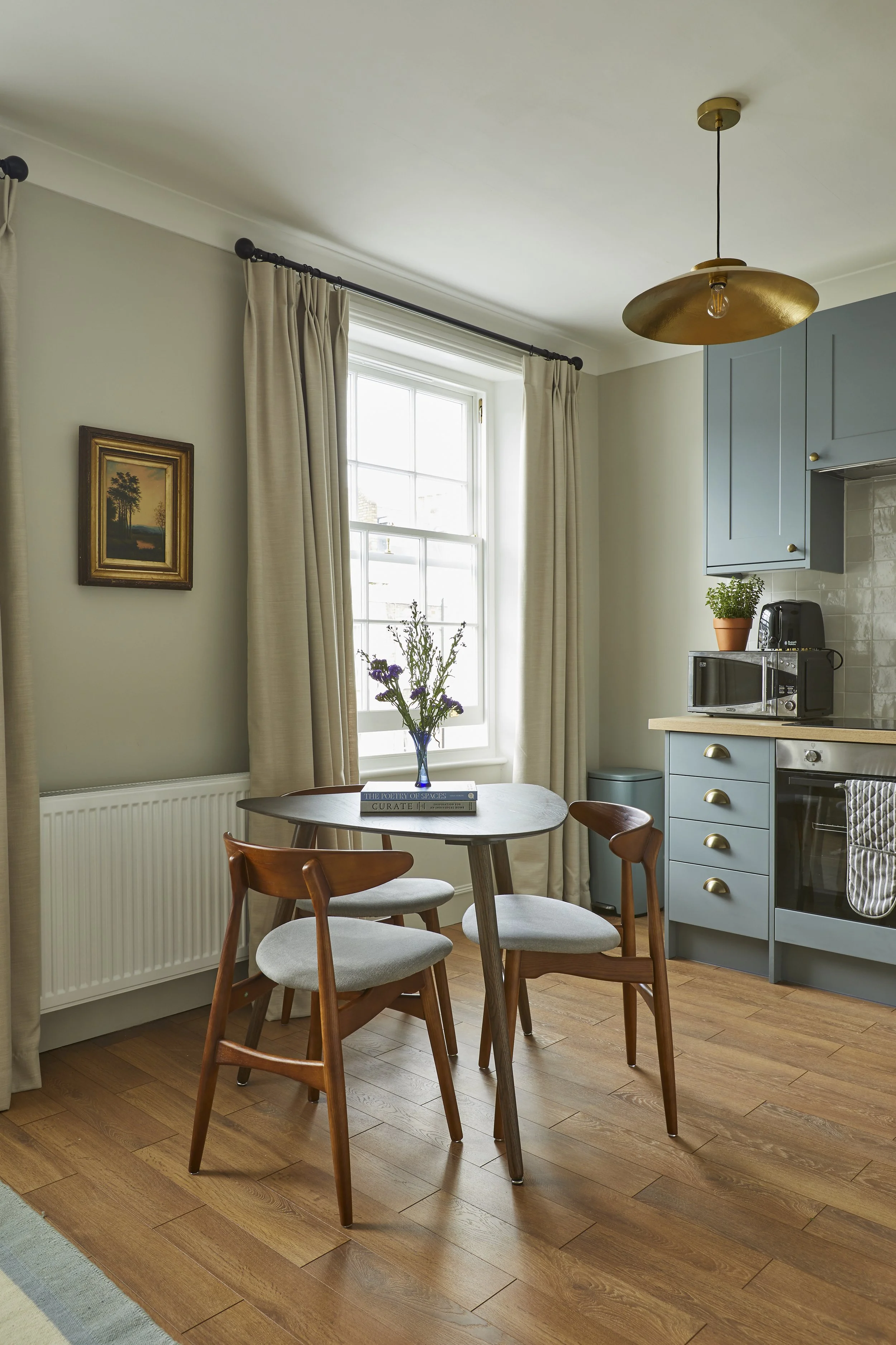 A cozy kitchen and dining area with a round wooden table, four wooden and upholstered chairs, a window with beige curtains, a blue kitchen cabinet, a small potted plant, a microwave, and a modern ceiling light fixture.