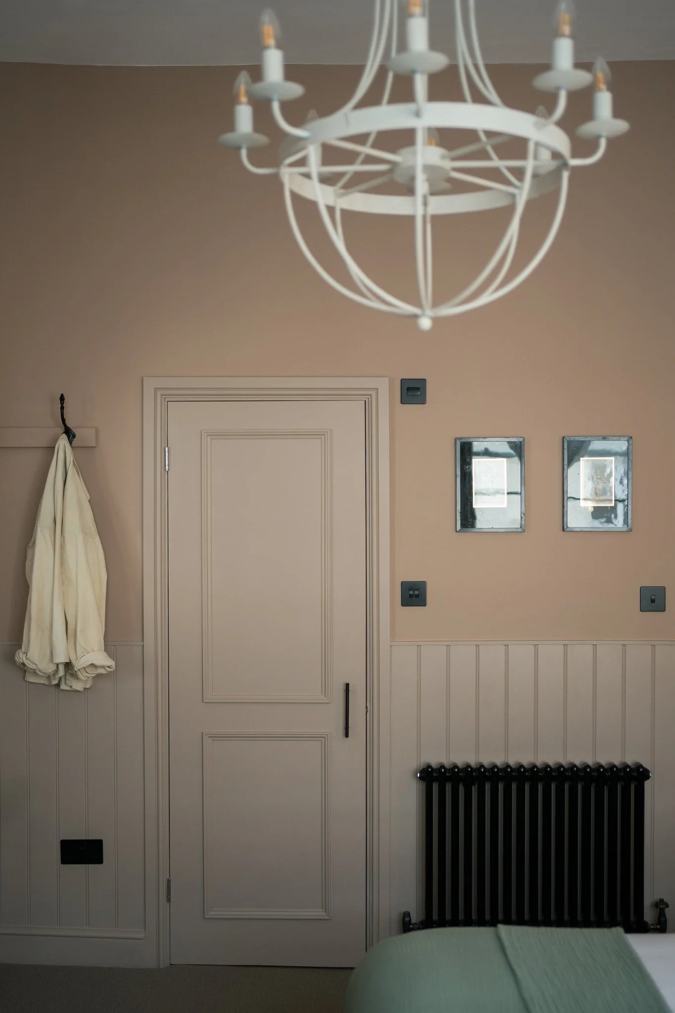 Interior room with beige walls, white door, black radiator, hanging white robe, two small windows, and a white chandelier.
