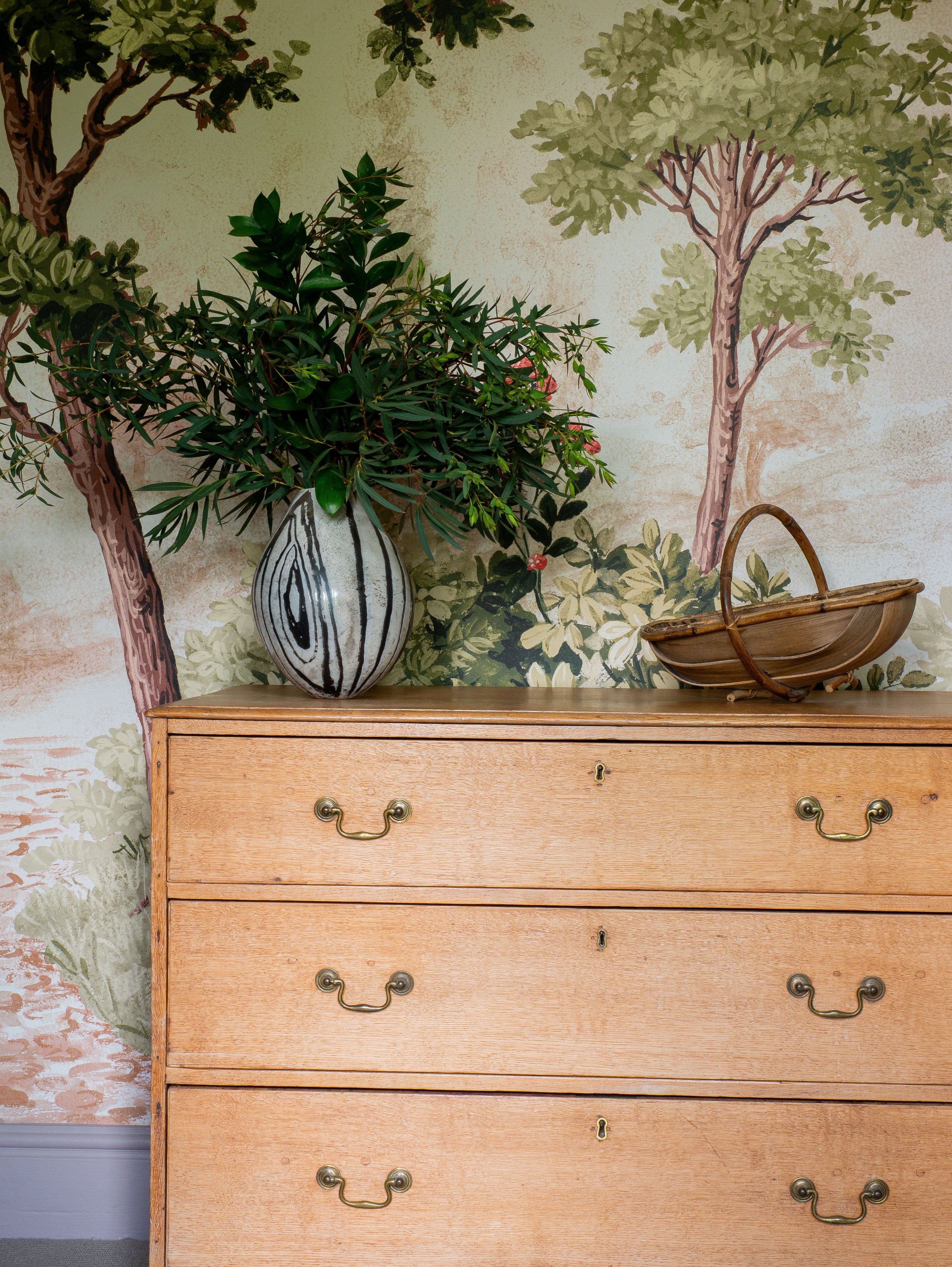 A wooden dresser with metallic handles, a decorative vase with green foliage and pink flowers, and a small wooden basket on top are set against a mural-style wallpaper depicting trees and foliage.