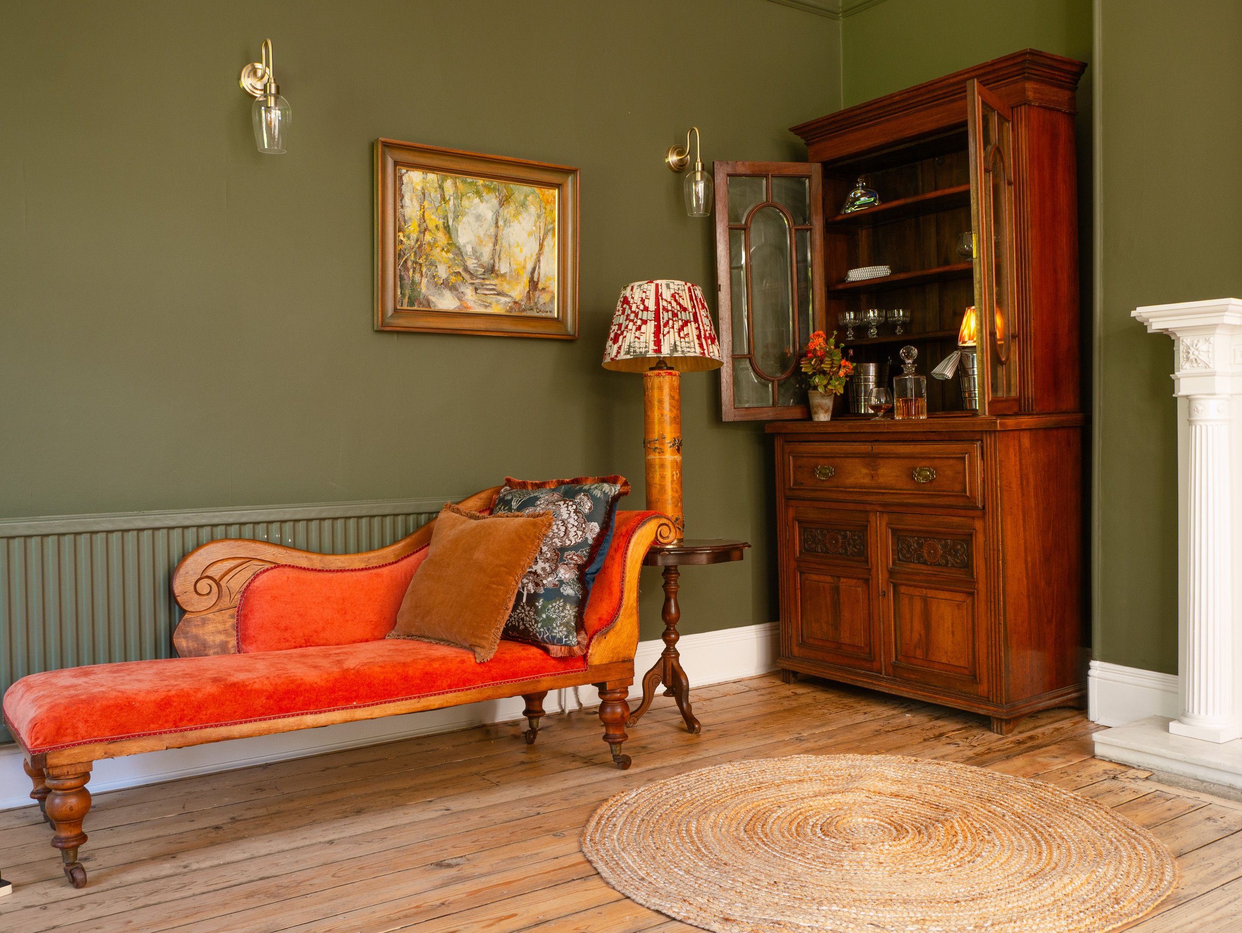 Living room with green walls, wooden furniture including a vintage chaise lounge with coral and patterned cushions, a wooden cabinet with glass doors, a framed landscape painting, a floral lamp, and a circular woven rug on hardwood floors.