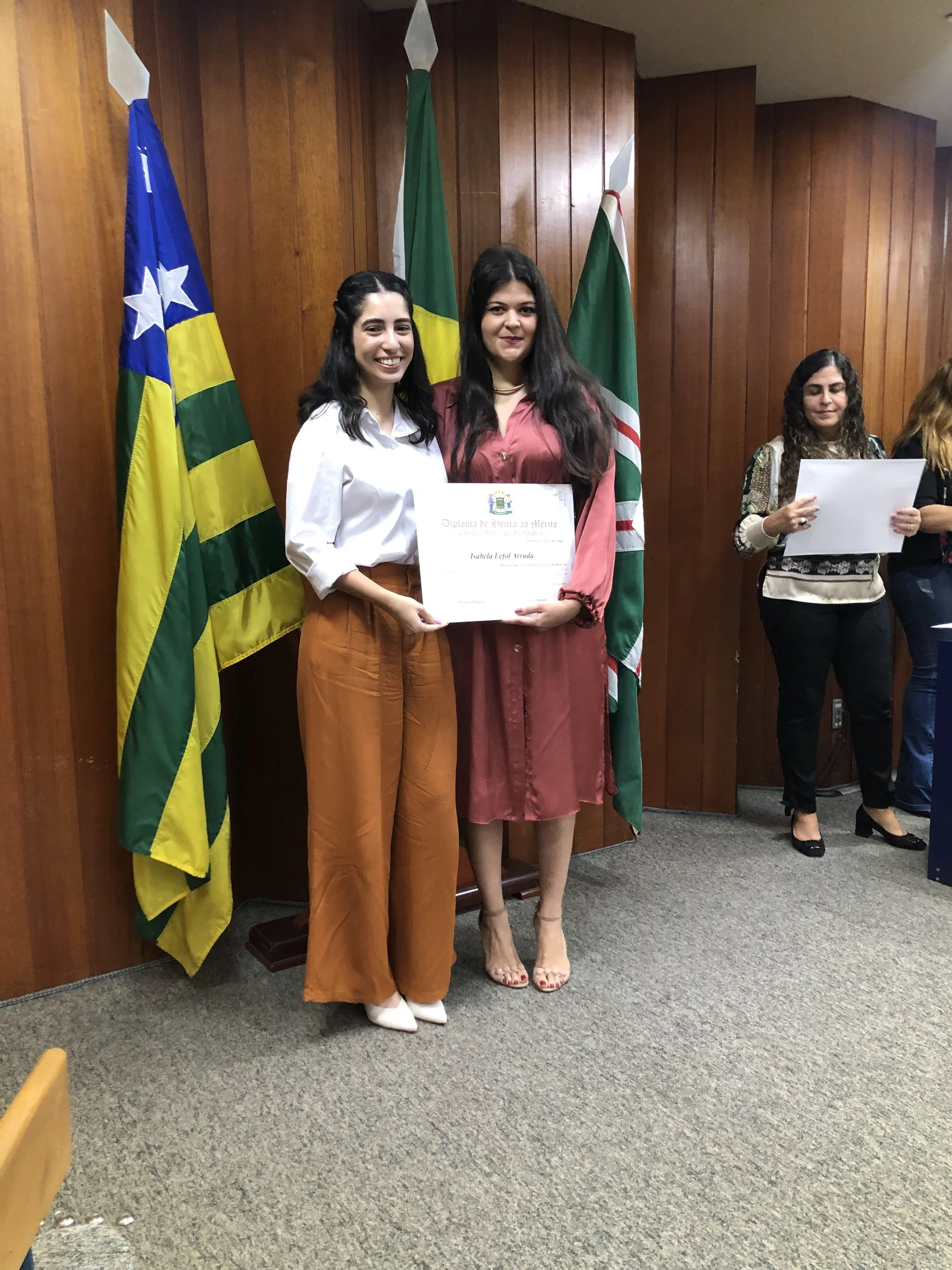 Two women standing in front of flags, holding a diploma, during an award ceremony. One woman wears a white blouse and brown pants, and the other wears a pink dress. Two women in the background hold certificates, in a room with wooden paneling.