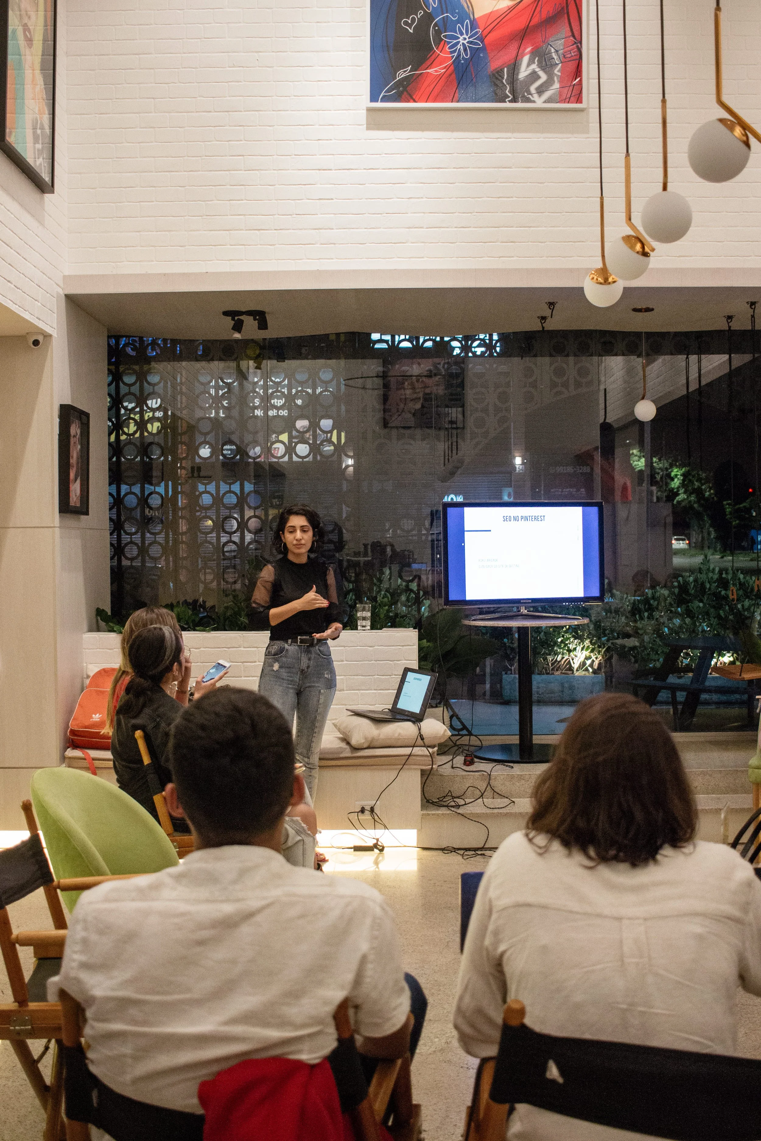 A woman giving a presentation to a small group of people in a modern, well-lit indoor space with artwork on the white brick walls and a large screen displaying a slide titled 'SEO NO PINTEREST.'